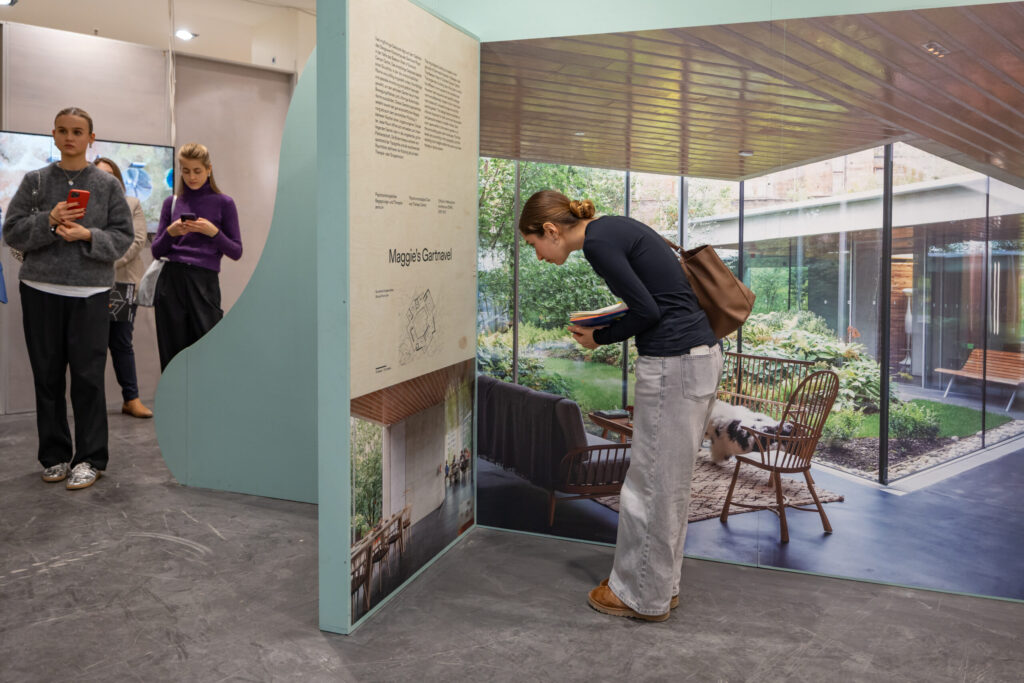 Eine Frau bückt sich, um ein Design-Display in einer Ausstellung genau zu betrachten, während zwei andere Frauen in der Nähe sind - die eine telefoniert, die andere schreibt - und das Display ein großes Foto eines gemütlichen Wohnzimmers mit Gartenblick zeigt. at Hochschule Coburg