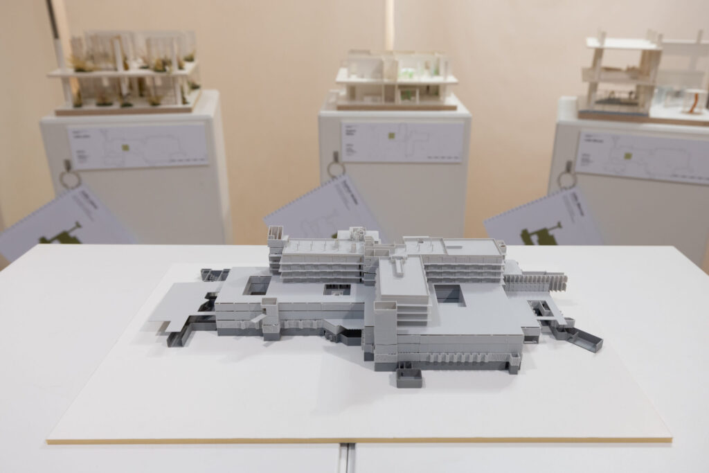 Ein detailliertes architektonisches Entwurfsmodell eines modernen Gebäudes ist auf einem Tisch ausgestellt, während im Hintergrund drei weitere Gebäudemodelle auf Sockeln und Diagrammen zu sehen sind. at Hochschule Coburg