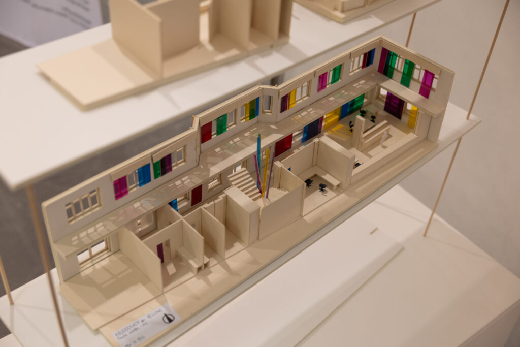 Ein detailliertes architektonisches Modell unterstreicht das durchdachte Design: zwei Stockwerke mit bunten Fensterelementen oben und beigen Zimmern und Fluren unten, die alle auf einer weißen Plattform stehen. at Hochschule Coburg