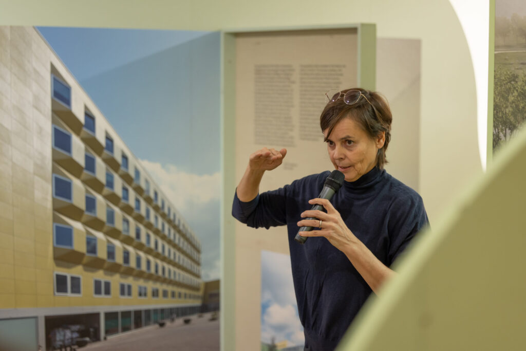 Eine Frau mit kurzen Haaren spricht in ein Mikrofon und gestikuliert mit einer Hand. Hinter ihr ist ein großes Foto zu sehen, das moderne Gebäude mit Balkonen und blauem Himmel zeigt. Sie trägt eine Brille auf dem Kopf und ein dunkles Oberteil. at Hochschule Coburg