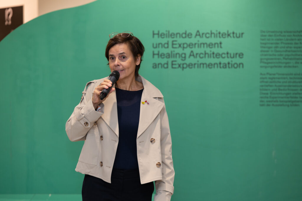Eine Frau in einer beigen Jacke spricht in ein Mikrofon vor einer grünen Wand mit der deutschen und englischen Aufschrift "Heilende Architektur und Experiment / Healing Architecture and Design Experimentation". at Hochschule Coburg