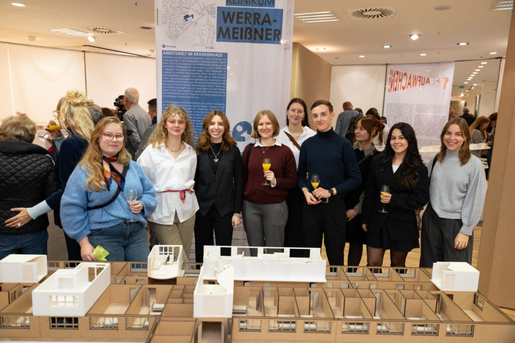 Eine Gruppe von neun lächelnden Menschen steht mit Getränken in der Hand hinter einem detaillierten Architekturmodell in einer Innenausstellung mit Informationsbannern und Displays im Hintergrund. at Hochschule Coburg