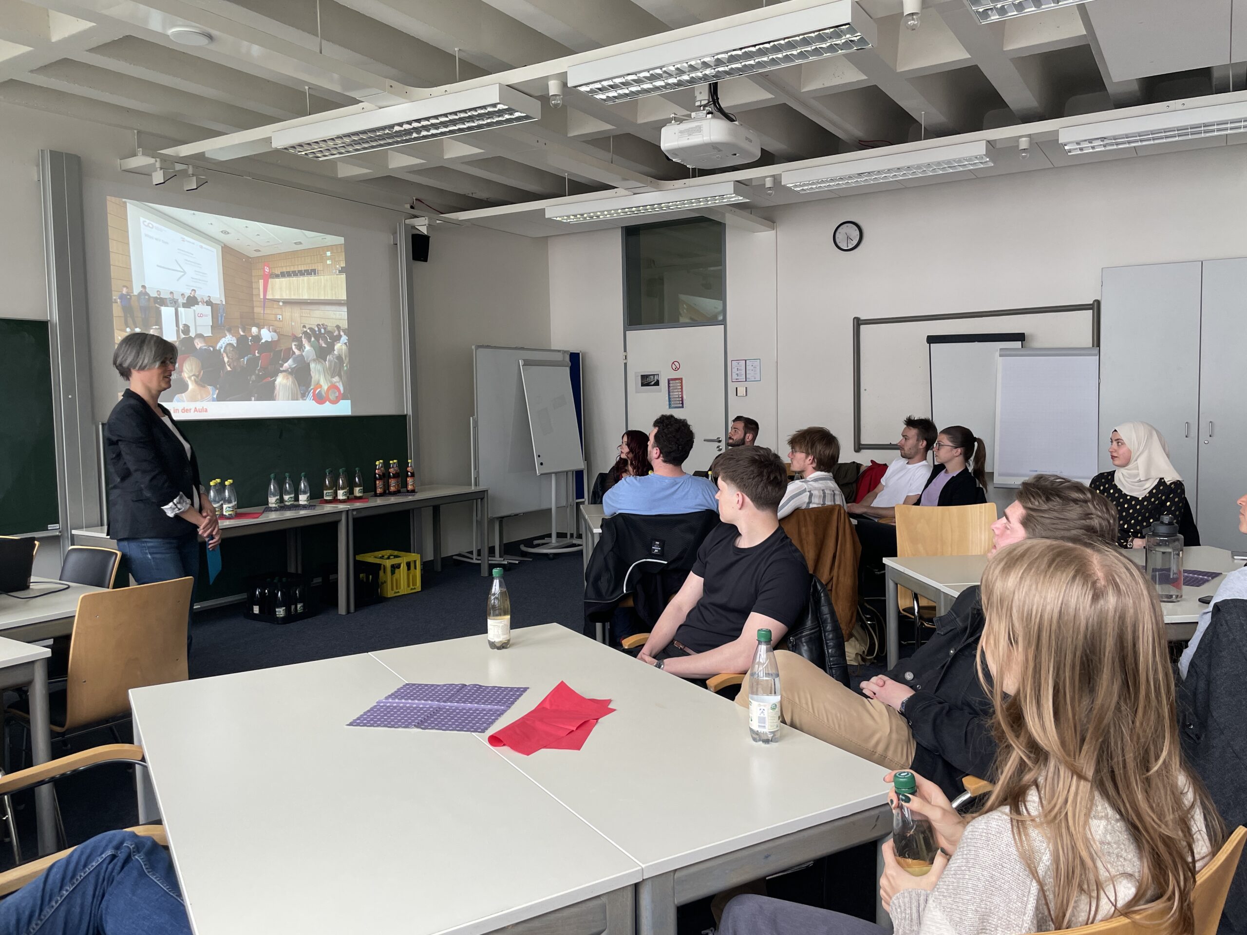 Eine Gruppe von Personen sitzt in einem Klassenzimmer um Tische herum und sieht zu, wie eine Frau einen auf eine Leinwand projizierten Vortrag über soziales Engagement hält. Auf den Tischen stehen Flaschen und Notizblöcke, und einige Teilnehmer halten Getränke in der Hand. at Hochschule Coburg