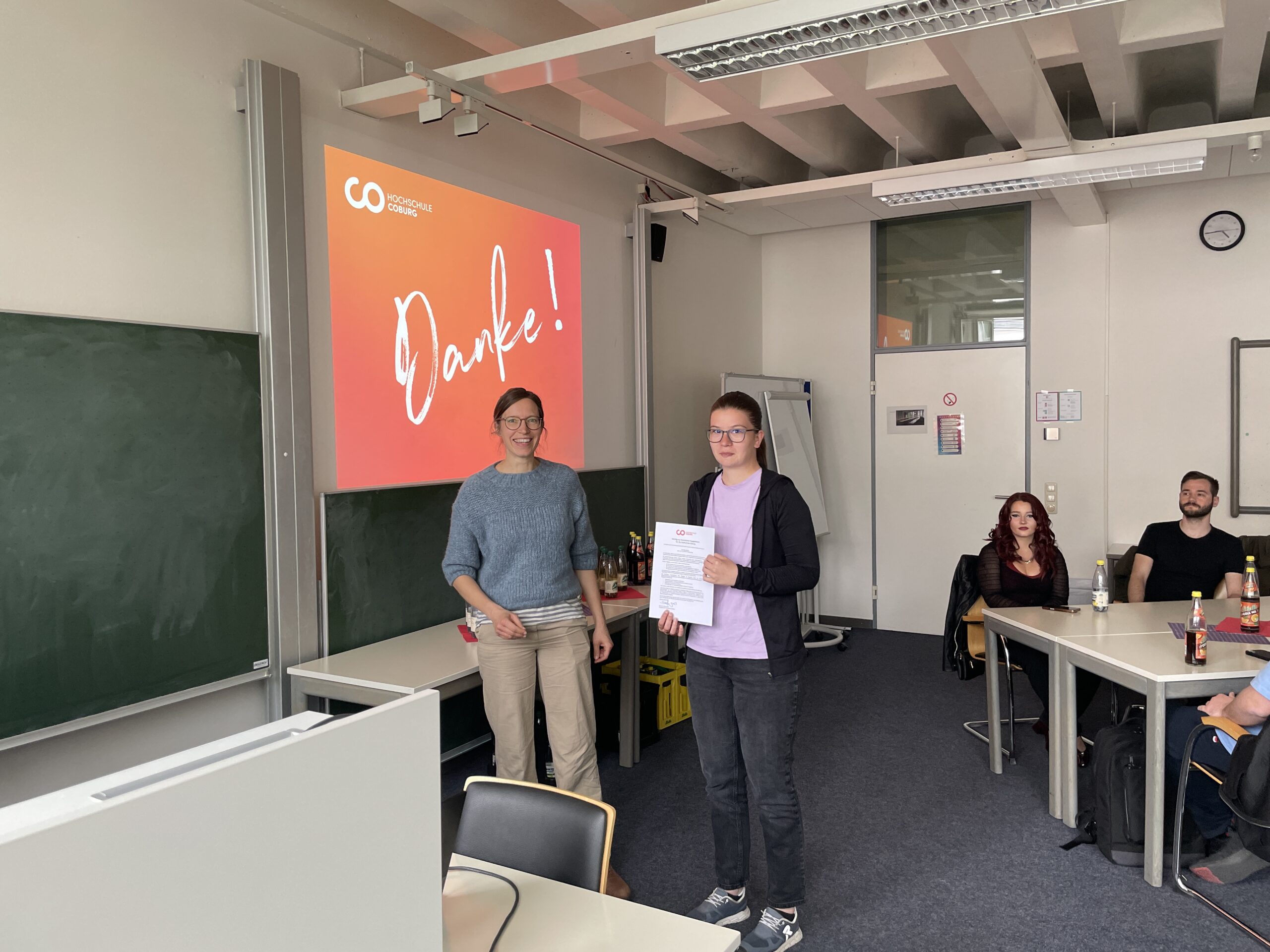 Zwei Personen stehen vorne in einem Klassenzimmer; die eine überreicht der anderen eine Urkunde, die Engagement für andere würdigt. Auf einer Leinwand hinter ihnen steht in großen Buchstaben "Danke!", während mehrere Personen an den Tischen die Szene beobachten. at Hochschule Coburg