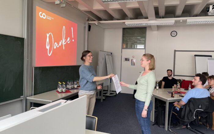 Zwei Frauen stehen vorne in einem Klassenzimmer und überreichen der anderen eine Urkunde für soziales Engagement. Ein Projektor zeigt "Danke!" auf der Leinwand an, während mehrere Personen sitzen und zusehen. at Hochschule Coburg