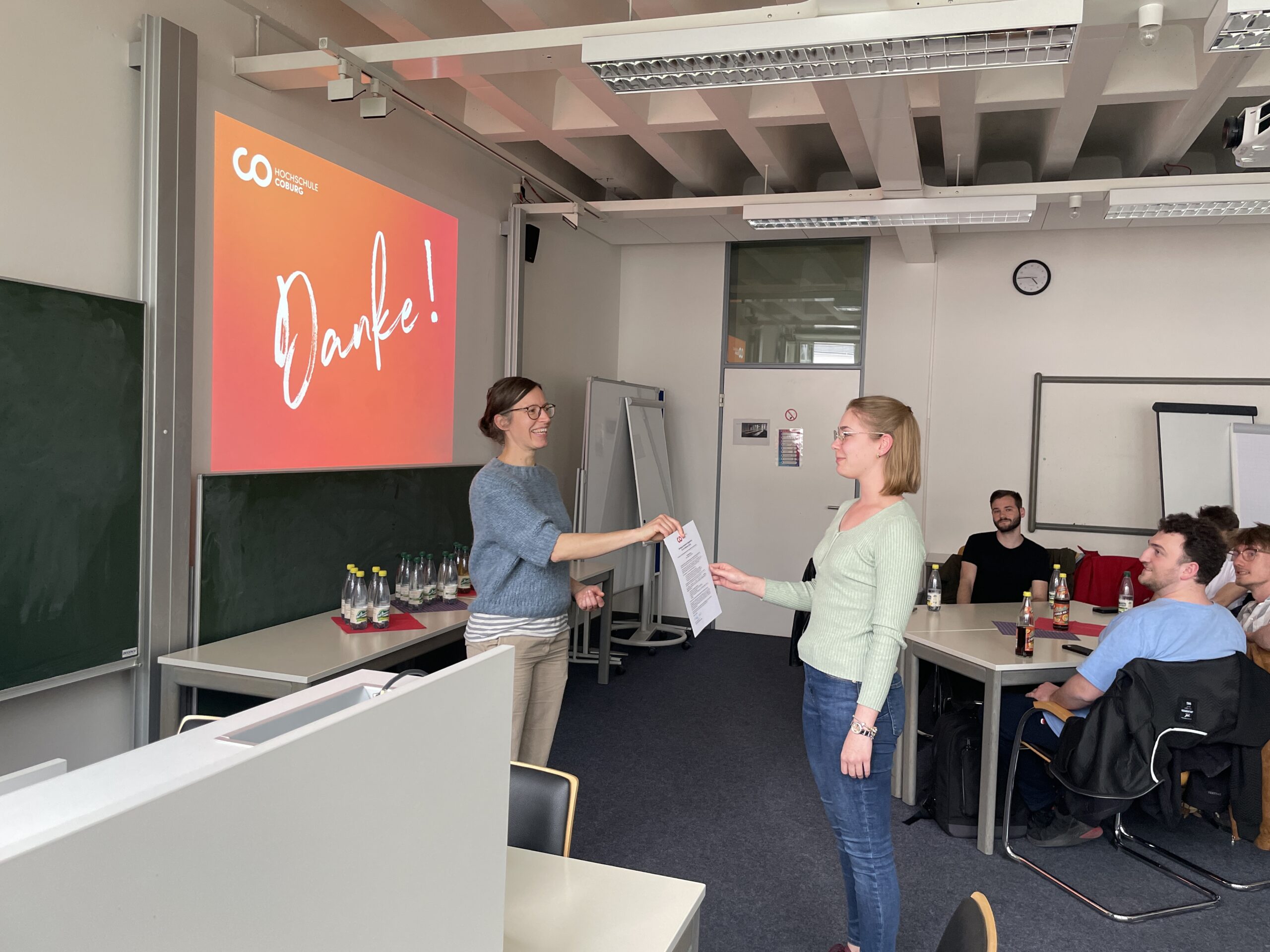 Zwei Frauen stehen vorne in einem Klassenzimmer und überreichen der anderen eine Urkunde für soziales Engagement. Ein Projektor zeigt "Danke!" auf der Leinwand an, während mehrere Personen sitzen und zusehen. at Hochschule Coburg