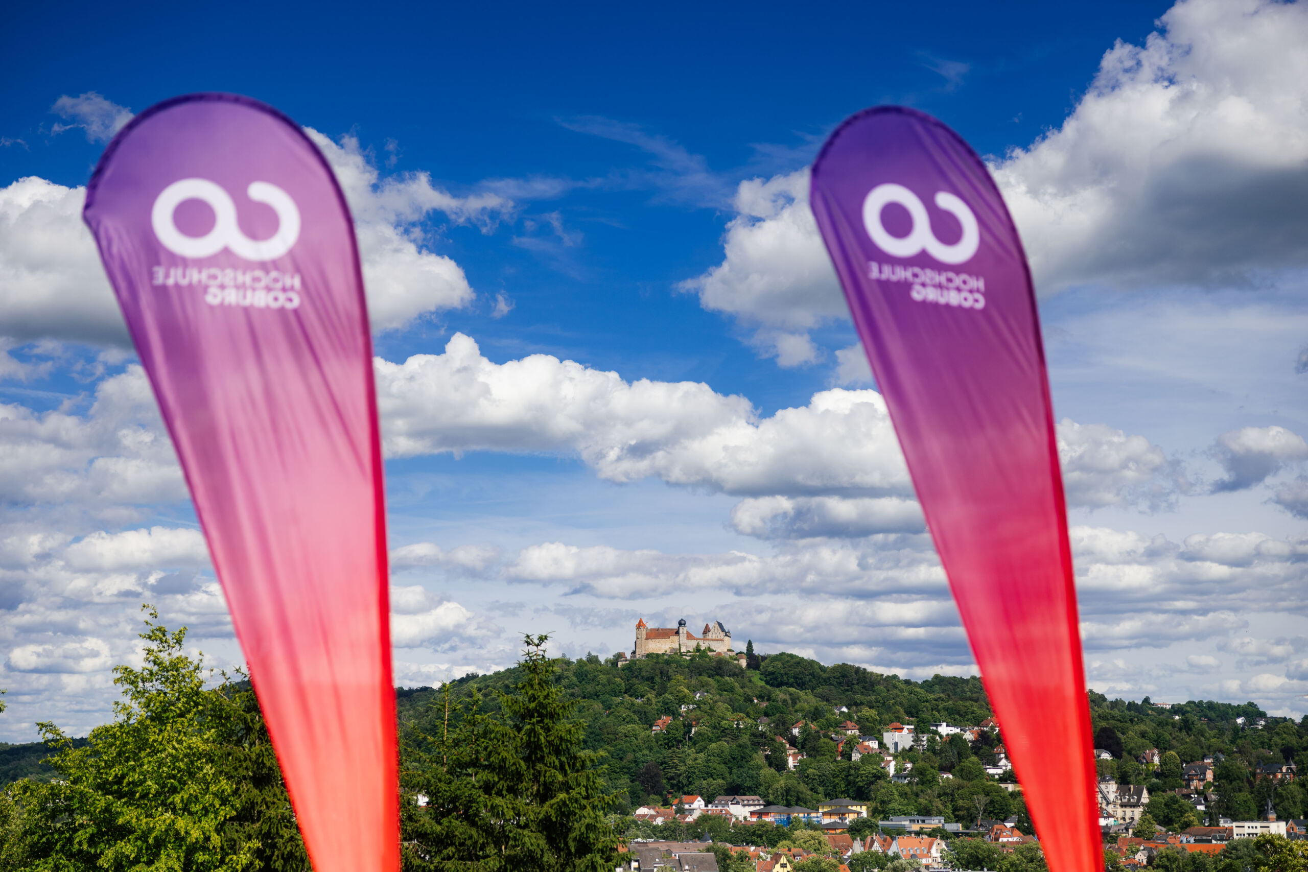 Im Vordergrund stehen zwei vertikale Federfahnen mit einem violetten und roten Farbverlauf und einem weißen Logo, die für „Wissenschaft“ werben; im Hintergrund ist eine Burg auf einem Hügel zu sehen, unter der sich eine Stadt unter blauem Himmel mit vereinzelten Wolken erstreckt. at Hochschule Coburg