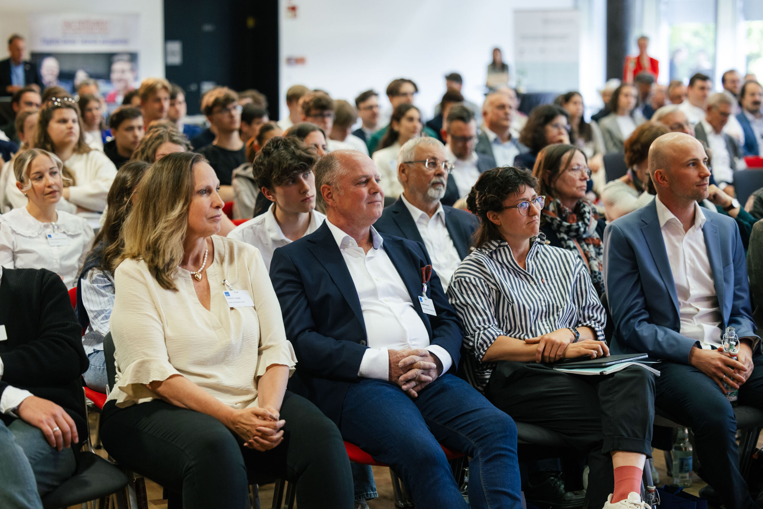 Eine bunt gemischte Gruppe von Menschen in Geschäftskleidung sitzt aufmerksam in Reihen bei einem Wirtschaftsseminar, blickt nach vorne und lauscht einem Redner. Das Publikum wirkt interessiert und konzentriert. at Hochschule Coburg