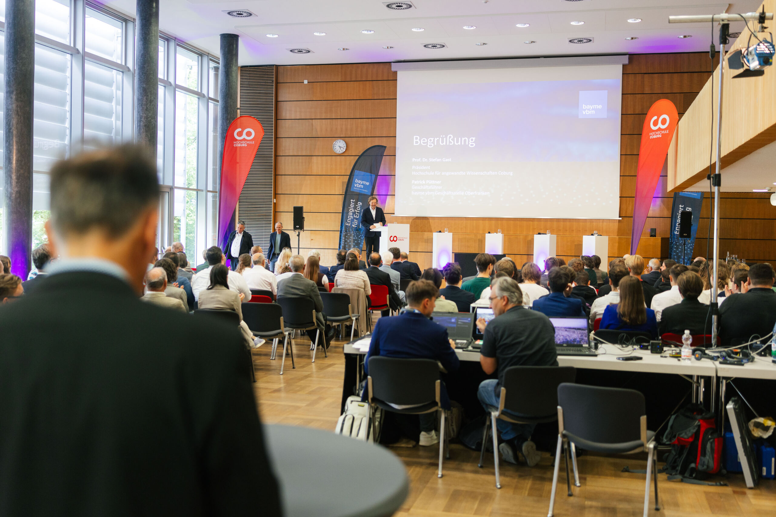 Ein Konferenzraum voller sitzender Teilnehmer, die auf eine Bühne blicken, auf der ein Redner an einem Rednerpult vor einer großen Leinwand steht, auf der „Begrüßung“ zu lesen ist. Rote Banner heben das Thema „Chancen durch Dialog“ zwischen Wissenschaft und Wirtschaft hervor. at Hochschule Coburg