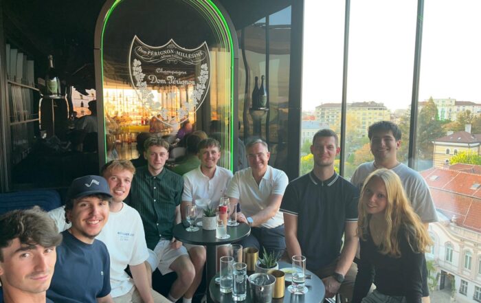 Eine Gruppe von neun jungen Erwachsenen sitzt lächelnd an zwei kleinen Tischen mit Getränken in einer modernen Bar mit einem Dom-Pérignon-Schild. Auf einem Bildschirm erscheint die Meldung „Automatisch gespeicherter Entwurf“, während durch große Fenster der Blick auf die Gebäude der Stadt bei Sonnenuntergang freigibt. at Hochschule Coburg