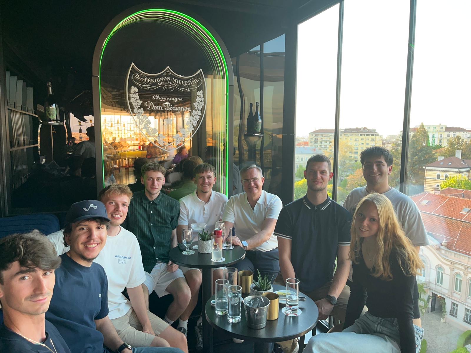 Eine Gruppe von neun jungen Erwachsenen sitzt lächelnd an zwei kleinen Tischen mit Getränken in einer modernen Bar mit einem Dom-Pérignon-Schild. Auf einem Bildschirm erscheint die Meldung „Automatisch gespeicherter Entwurf“, während durch große Fenster der Blick auf die Gebäude der Stadt bei Sonnenuntergang freigibt. at Hochschule Coburg