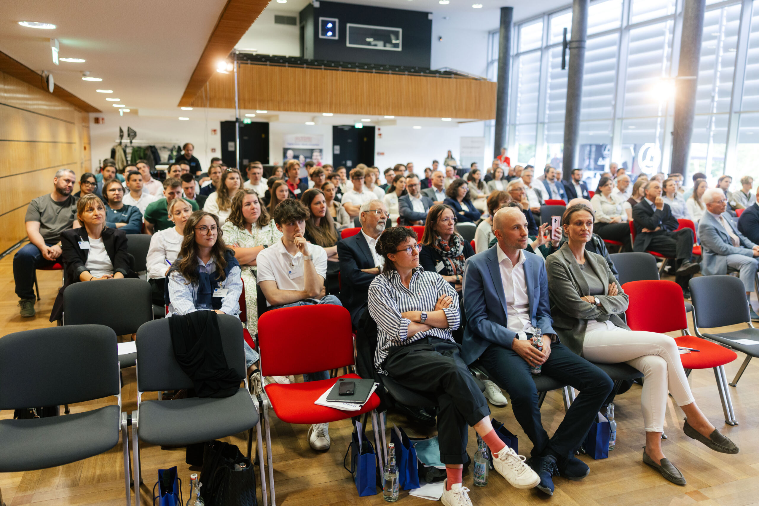 Eine große Gruppe von Menschen sitzt in Reihen roter Stühle in einem hellen Konferenzraum und hört aufmerksam einem Vortrag zum Thema Wirtschaft zu. Einige Teilnehmer scheinen sich intensiv am Dialog zu beteiligen, während andere entspannt oder nachdenklich wirken. at Hochschule Coburg