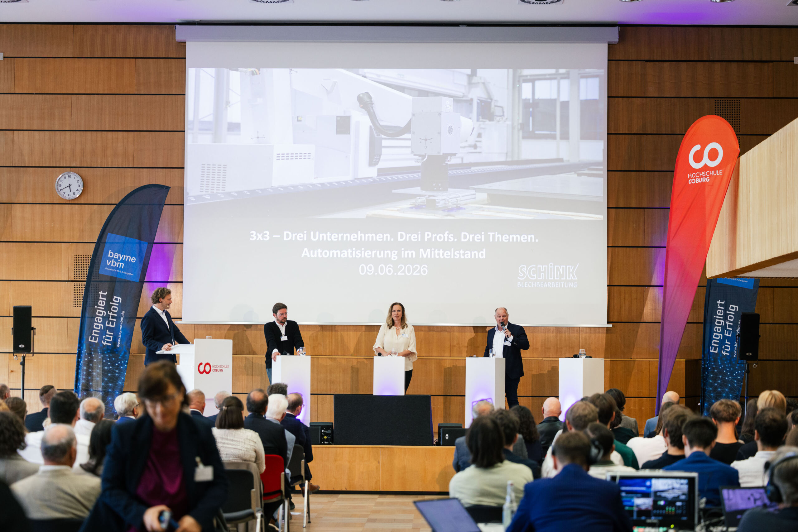 Auf der Bühne steht eine Diskussionsrunde aus vier Referenten, die mit dem Publikum ins Gespräch kommen, während auf einer großen Leinwand hinter ihnen Informationen zur Automatisierung in mittelständischen Unternehmen gezeigt werden. Die Veranstaltung findet in einem modernen Auditorium statt, was den Schwerpunkt auf Wirtschaft und Wissenschaft unterstreicht. at Hochschule Coburg
