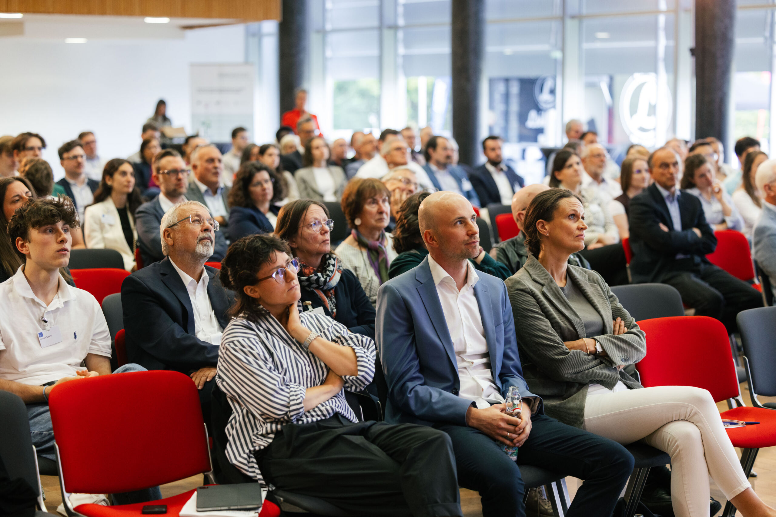 Eine bunt gemischte Gruppe von Menschen in Business- und Freizeitkleidung sitzt aufmerksam in Reihen roter Stühle bei einer Konferenz in einem Saal und nimmt am „Dialog über Wissenschaft und Wirtschaft“ teil, wobei ihr Blick auf einen Vortrag gerichtet ist, der außerhalb des Bildausschnitts stattfindet. at Hochschule Coburg