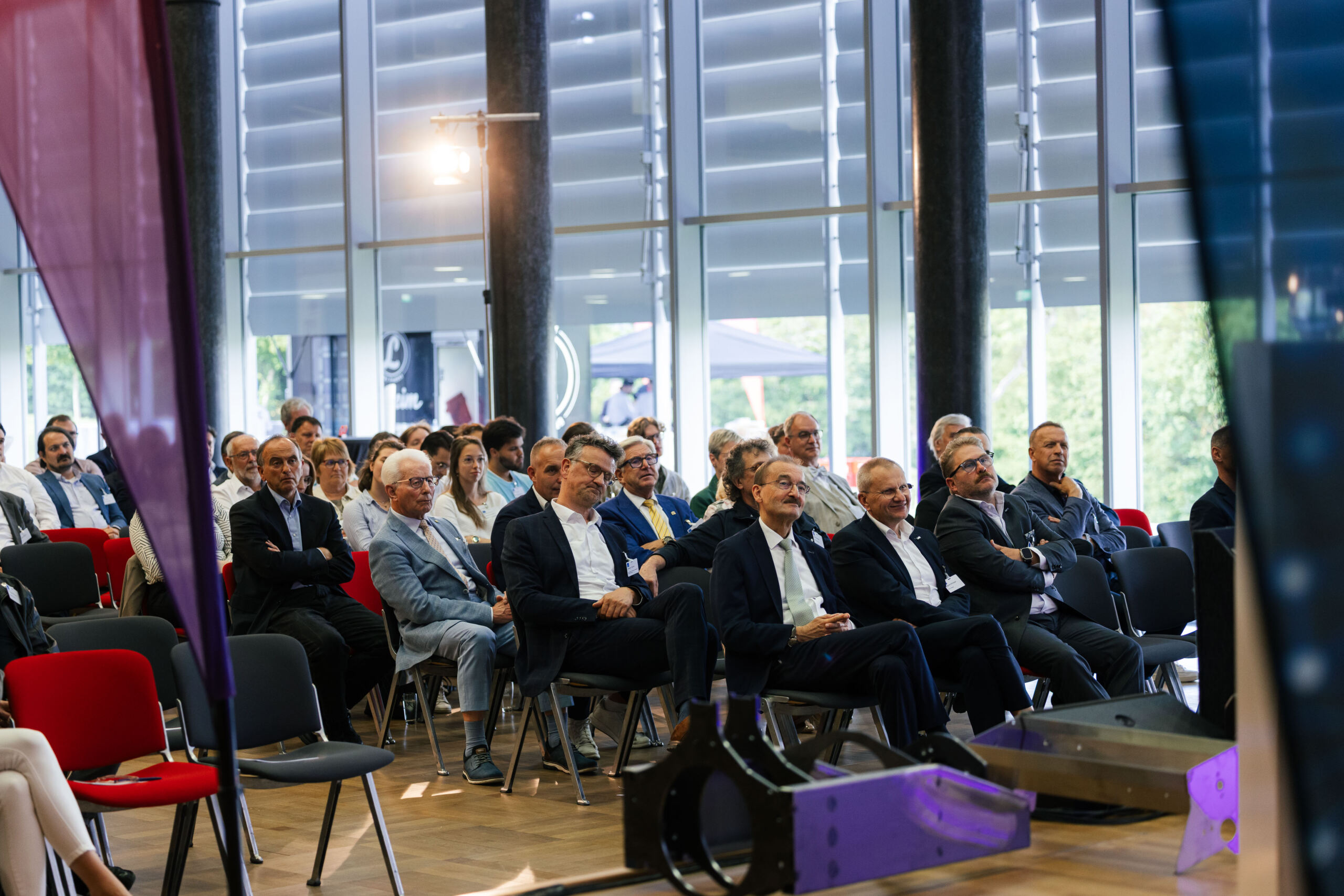 Eine Gruppe von Menschen in Geschäftskleidung sitzt aufmerksam in Stuhlreihen bei einer Wirtschaftskonferenz oder einem Seminar in einem Saal und ist in einen Dialog vertieft, im Hintergrund sind große Fenster und Tageslicht zu sehen. at Hochschule Coburg