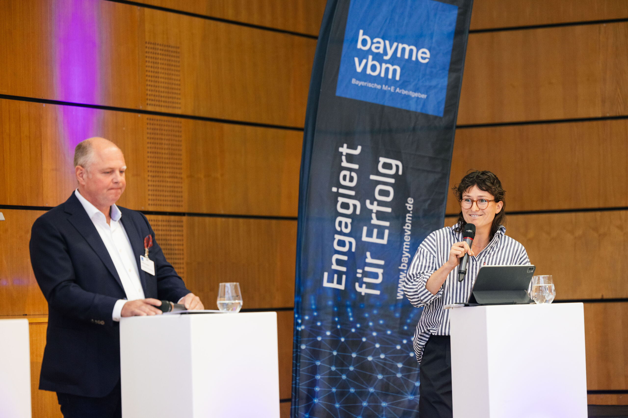 Zwei Personen sprechen an Rednerpult vor einem blauen Banner mit der deutschen Aufschrift „bayme vbm – Engagiert für Erfolg“ in einem modernen, mit Holz vertäfelten Raum und führen einen Dialog über Wirtschaft. Beide haben Mikrofone und Gläser mit Wasser vor sich stehen. at Hochschule Coburg