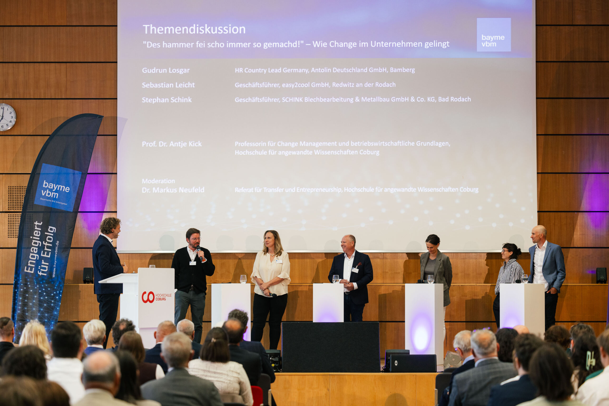 Auf der Bühne steht eine Diskussionsrunde aus sechs Referenten vor einem sitzenden Publikum und führt einen Dialog über Wissenschaft und Wirtschaft, wobei das Thema auf einer großen Leinwand hinter ihnen im Rahmen einer Fachkonferenz angezeigt wird. Auf der Bühne sind Banner und Rednerpulte zu sehen. at Hochschule Coburg