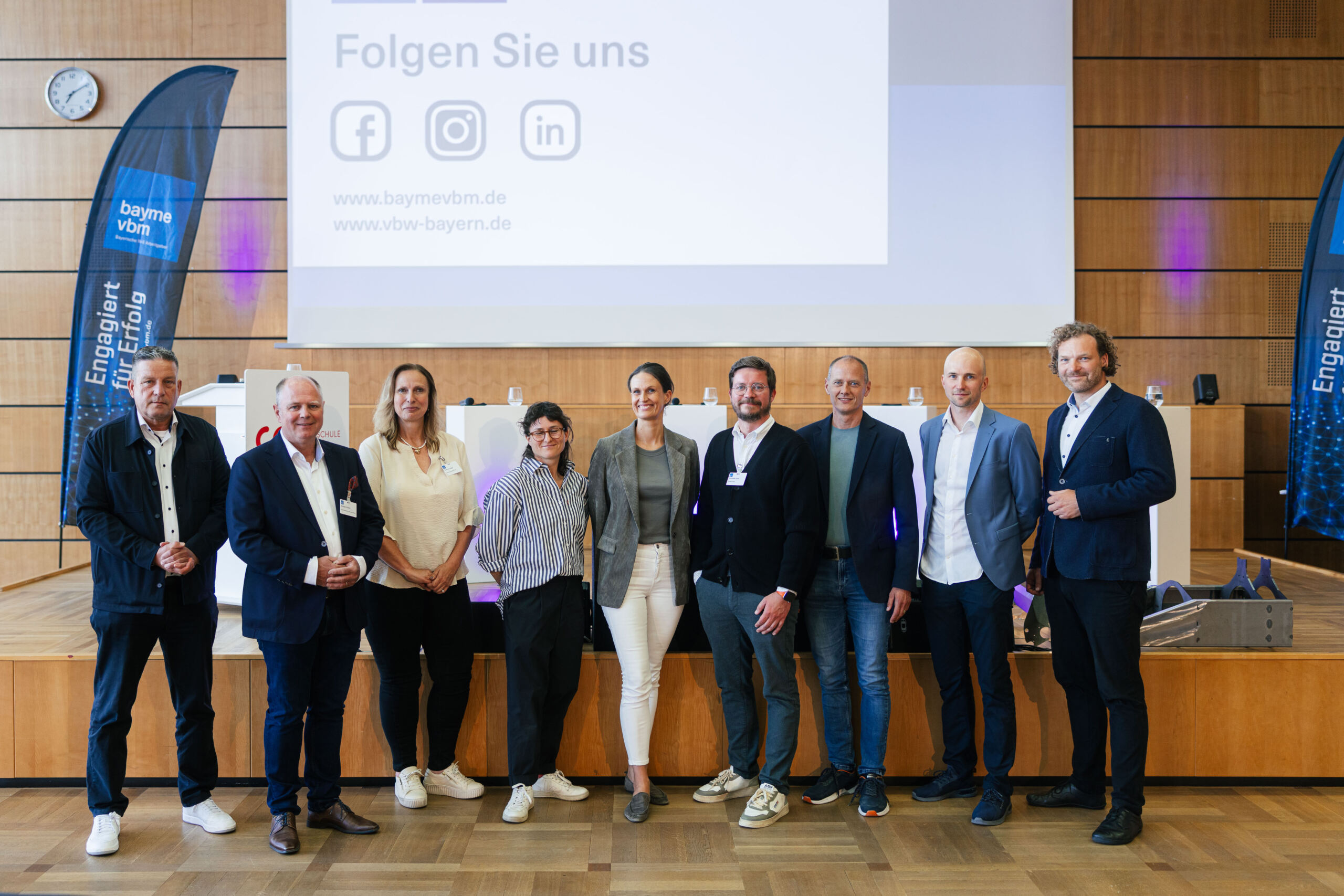Neun Personen stehen in einer Reihe auf einer Bühne in einem Saal, gekleidet in Business-Kleidung, hinter ihnen sind Banner und eine große Leinwand zu sehen, auf der Social-Media-Symbole und deutscher Text zu sehen sind – damit ist die Kulisse für einen spannenden Dialog zwischen Wissenschaft und Wirtschaft geschaffen. at Hochschule Coburg