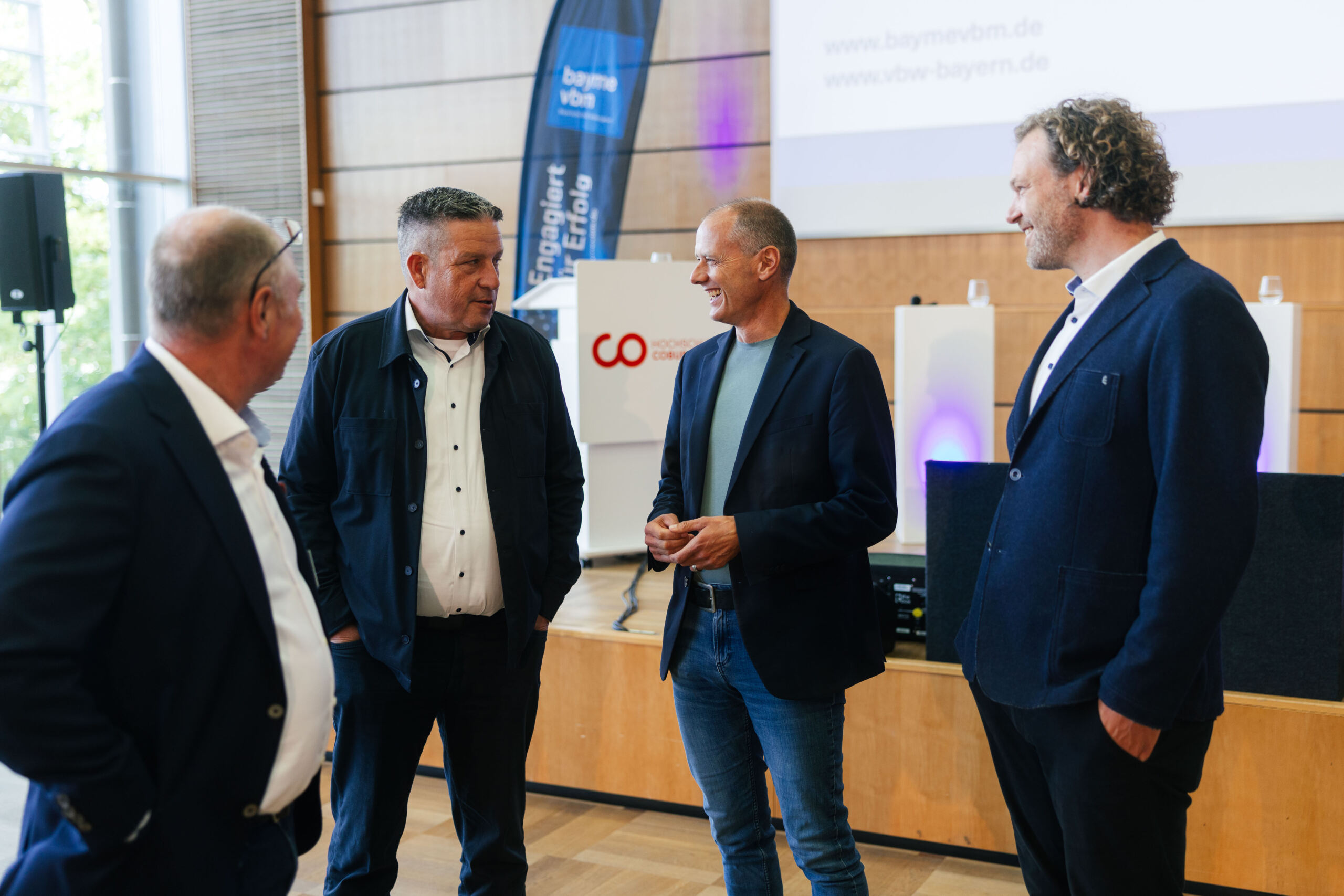 Vier Männer in Geschäftskleidung stehen in einem modernen Konferenzraum, führen ein lebhaftes Gespräch und lächeln. Im Hintergrund sind Präsentationsbildschirme und Banner zu sehen, auf denen die Begriffe „Wissenschaft“ und „Wirtschaft“ hervorgehoben sind. at Hochschule Coburg