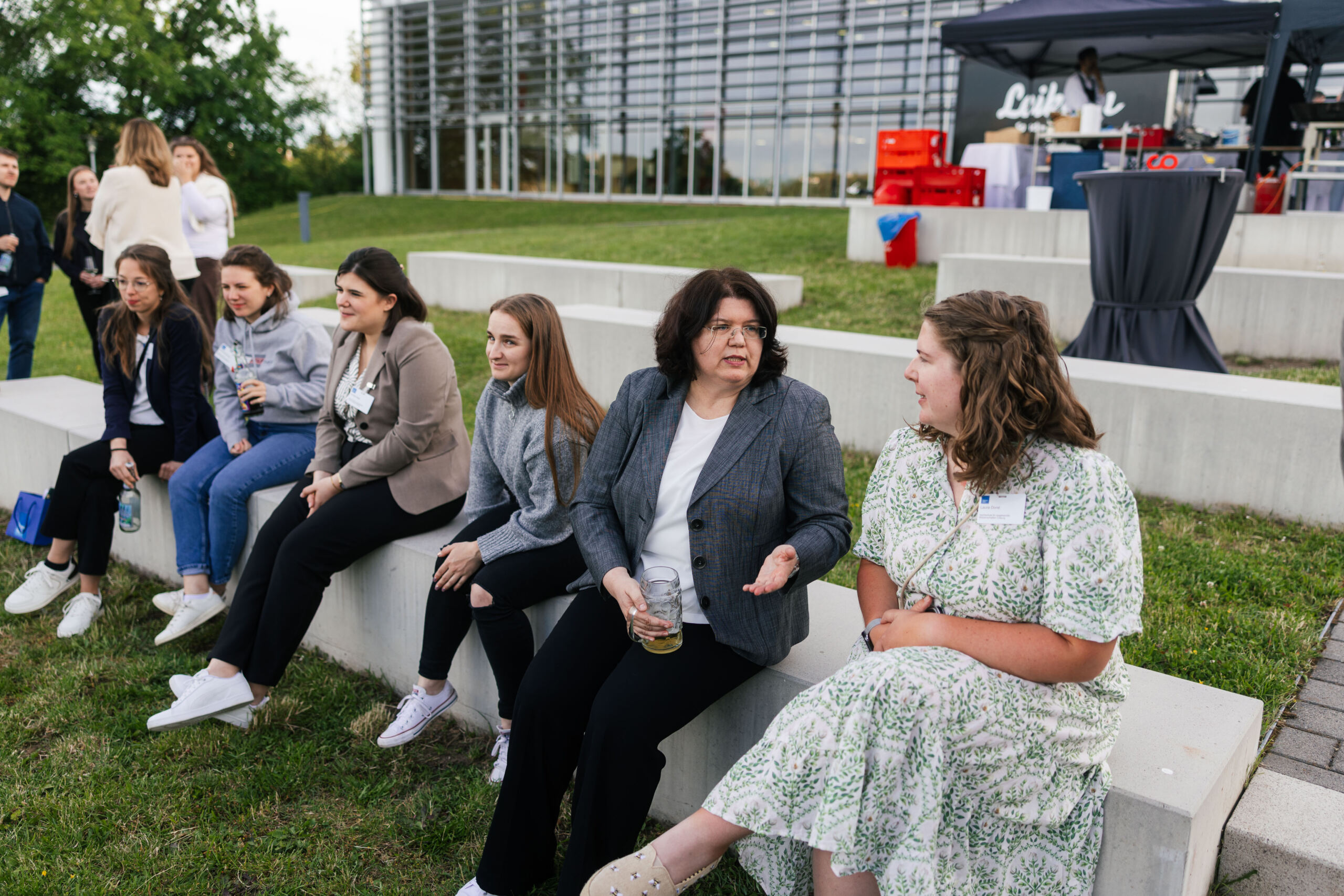 Eine Gruppe von Frauen sitzt auf Betonstufen im Freien in der Nähe eines modernen Glasgebäudes und unterhält sich, während sie Getränke in der Hand halten. Diese ungezwungene, gesellige Szene spielt sich vor dem Hintergrund eines 3x3-Getränkestands ab, der Menschen sowohl aus Wirtschaftskreisen als auch aus Wissenschaftskreisen anzieht. at Hochschule Coburg