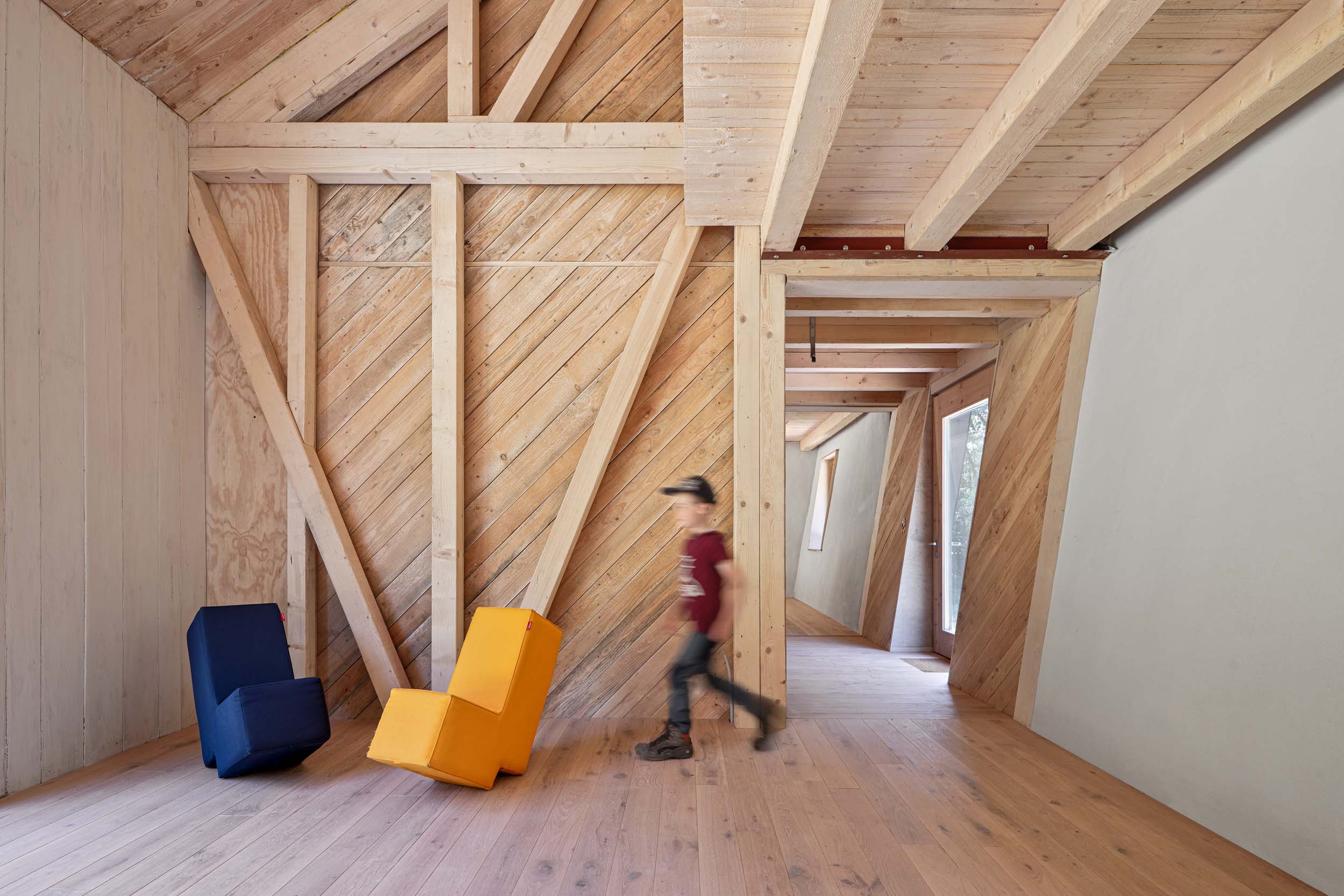 Ein Kind läuft durch einen modernen Raum im Waldkindergarten Pinzberg, der mit schrägen Holzwänden und Deckenbalken ausgestattet ist. Zwei farbenfrohe, minimalistische Stühle – blau und gelb – heben sich deutlich ab, während natürliches Licht hereinströmt und den Raum für die „Architektouren 2026“ in Szene setzt. at Hochschule Coburg