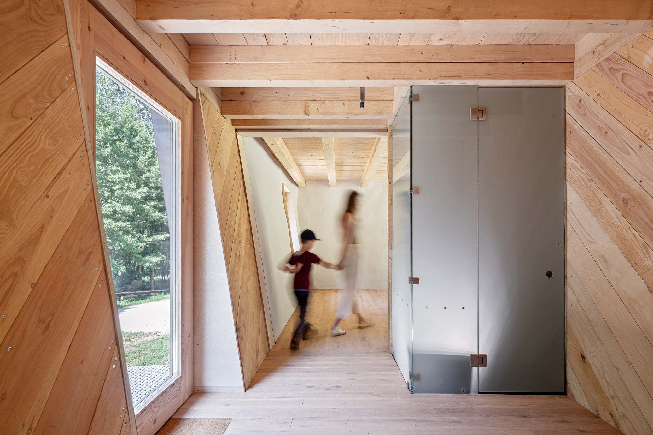 Ein modernes Holzinterieur mit großen Fenstern, schräg verlaufenden Holzvertäfelungen und einer Glastür. Ein Erwachsener und ein Kind gehen Hand in Hand den hellen Flur des Waldkindergartens Pinzberg entlang, während draußen üppiges Grün zu sehen ist – ein einladender Zwischenstopp bei den Architektouren 2026. at Hochschule Coburg