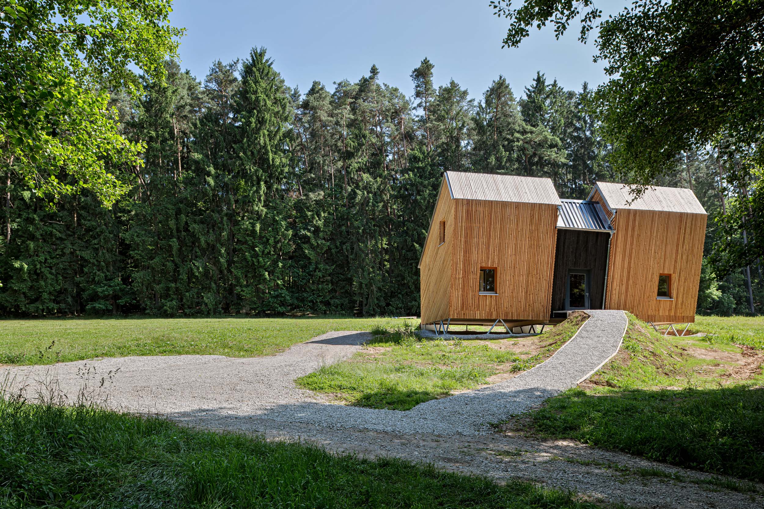 Ein modernes Holzhaus mit zwei Satteldächern auf einem grasbewachsenen Grundstück in der Nähe eines dichten Waldes, entworfen für den Waldkindergarten Pinzberg. Ein geschwungener Kiesweg führt zu einer Rampe hinauf zum Eingang, während Sonnenlicht durch die Bäume fällt – Teil der Architektouren 2026. at Hochschule Coburg