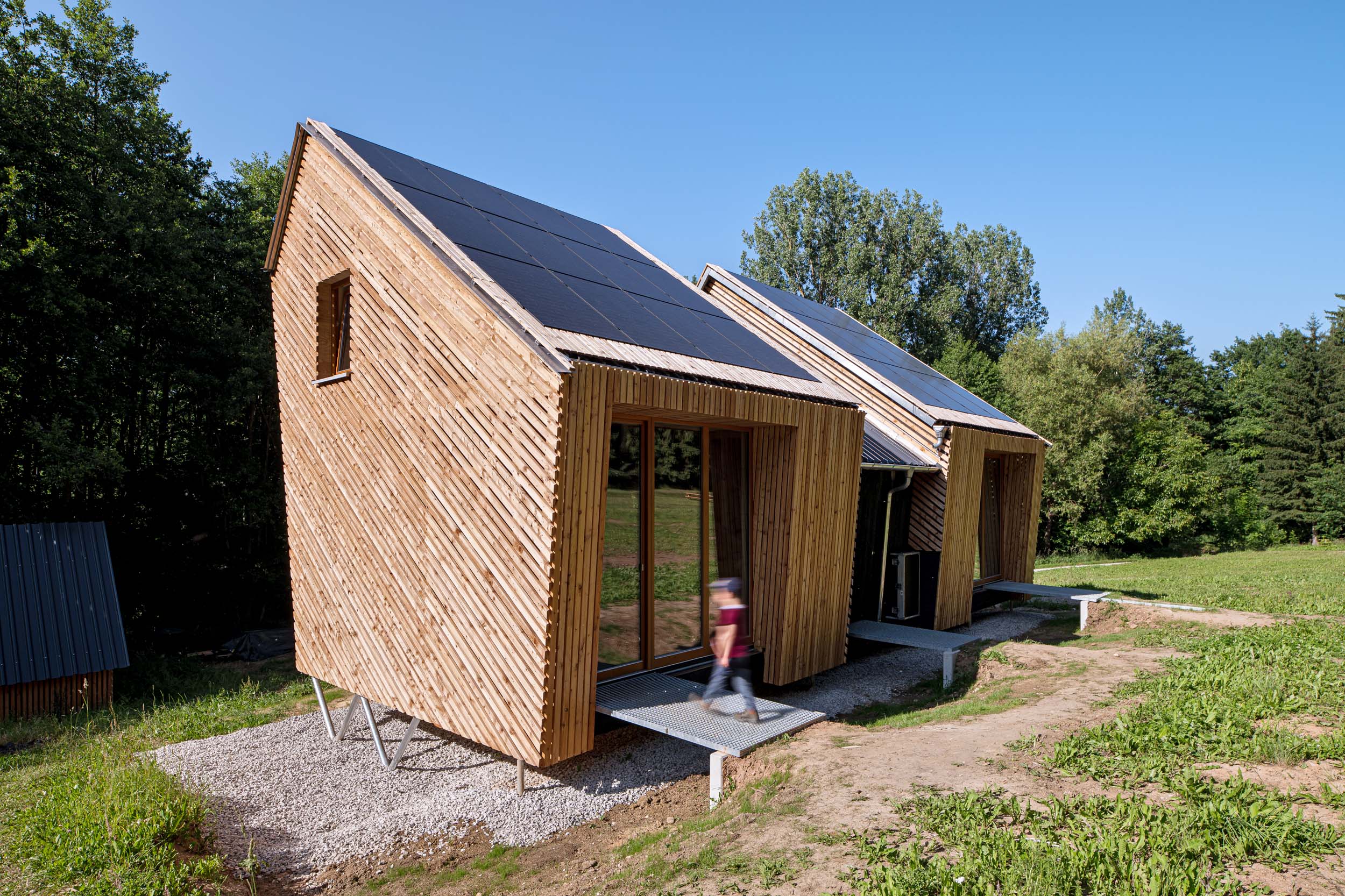 Zwei kleine, moderne Holzhütten mit schrägen, mit Solarmodulen bedeckten Dächern stehen auf einer Rasenfläche in der Nähe des Waldes und gehören zum Waldkindergarten Pinzberg. Ein Kind geht den Weg vor diesen minimalistischen Hütten entlang – ein idealer Anblick für die Architektouren 2026. at Hochschule Coburg