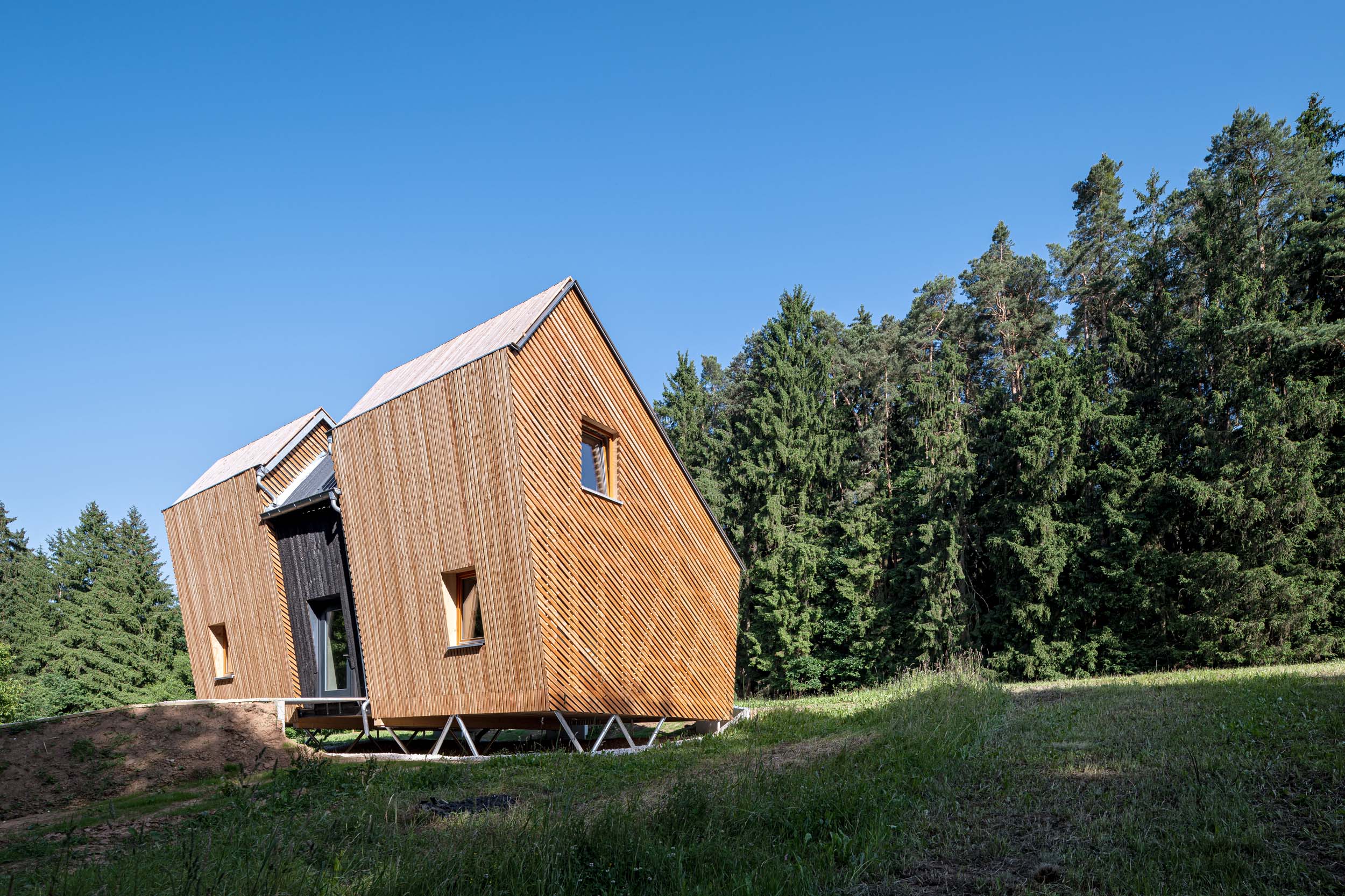 Ein modernes Holzhaus mit einem eckigen, schrägen Dach steht auf Stelzen auf einer grasbewachsenen Lichtung neben dem dichten Wald, der den Waldkindergarten Pinzberg umgibt, und präsentiert sich im Rahmen der „Architektouren 2026“ unter strahlend blauem Himmel. at Hochschule Coburg