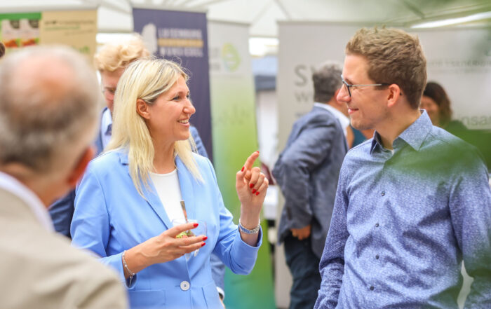 Eine Frau in einem hellblauen Blazer lächelt und gestikuliert, während sie sich auf dem Innovationskongress Oberfranken mit einem Mann mit Brille und einem blau gemusterten Hemd unterhält; im Hintergrund sind weitere Personen und Informationsstände zu sehen. at Hochschule Coburg