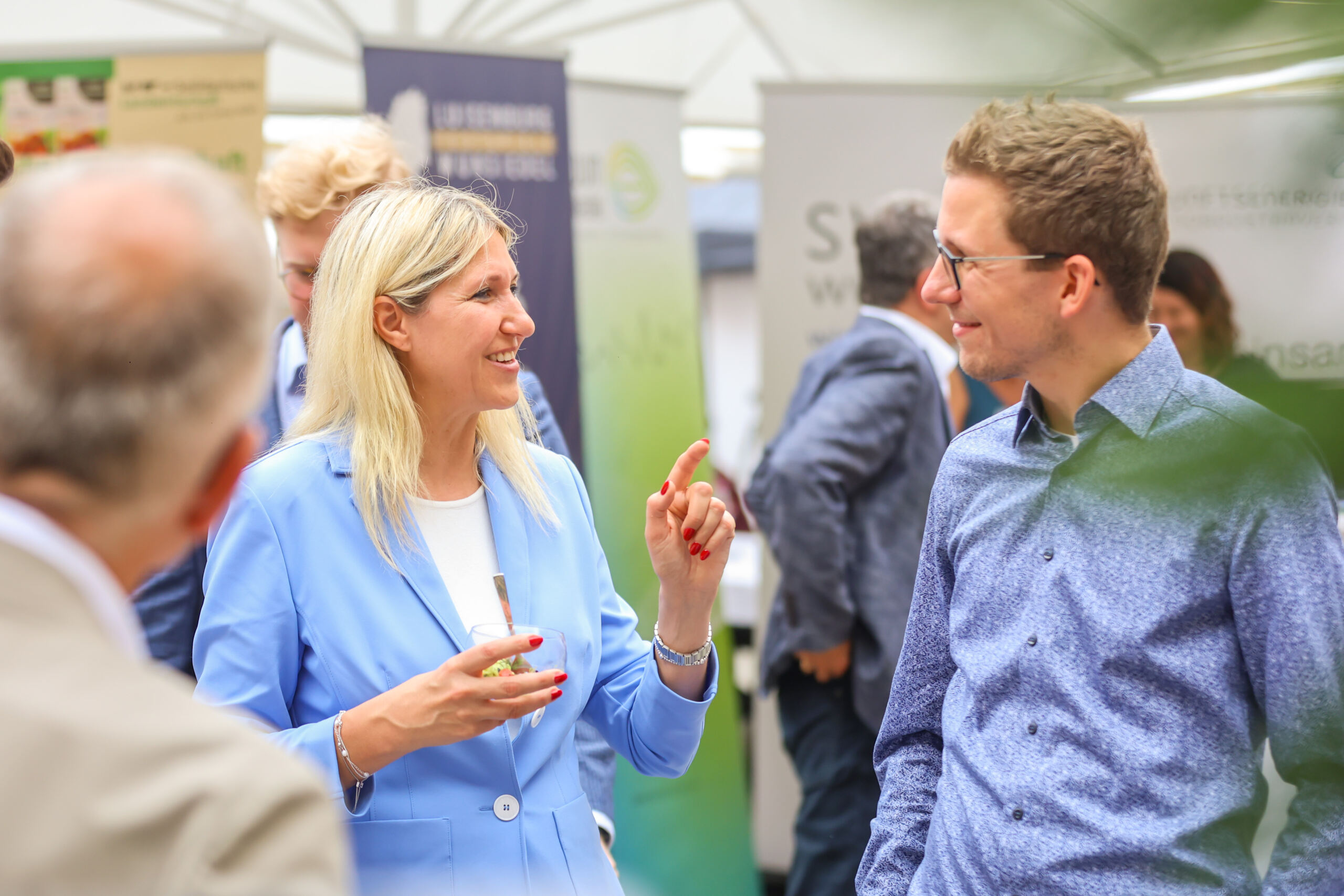 Eine Frau in einem hellblauen Blazer lächelt und gestikuliert, während sie sich auf dem Innovationskongress Oberfranken mit einem Mann mit Brille und einem blau gemusterten Hemd unterhält; im Hintergrund sind weitere Personen und Informationsstände zu sehen. at Hochschule Coburg