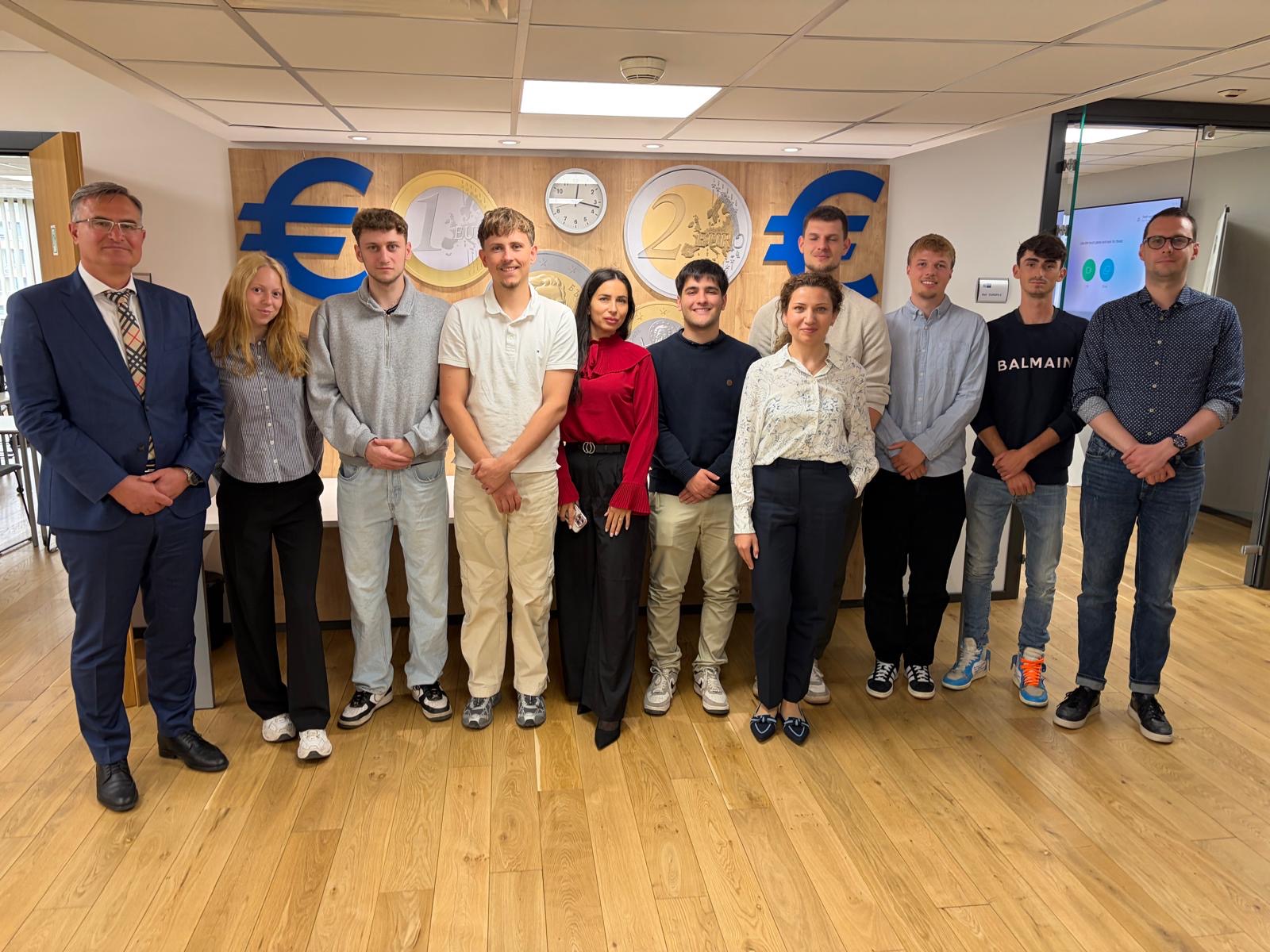 Eine Gruppe von elf Personen, die elegante und legere Kleidung tragen, steht in einem Raum auf einem Holzboden, hinter ihnen sind Euro-Scheine und Wanduhren zu sehen, und posiert für ein Gruppenfoto – vielleicht für einen automatisch gespeicherten Entwurf. at Hochschule Coburg