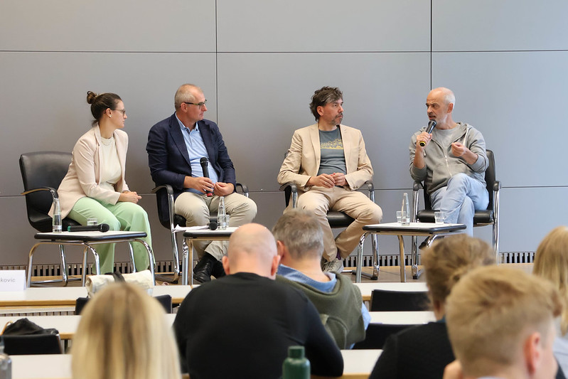 Vier Personen sitzen auf der Bühne und führen vor Publikum eine Podiumsdiskussion. Ein Mann auf der rechten Seite spricht in ein Mikrofon, während die anderen zuhören. Der Schauplatz scheint ein moderner Konferenzraum oder Hörsaal zu sein. at Hochschule Coburg