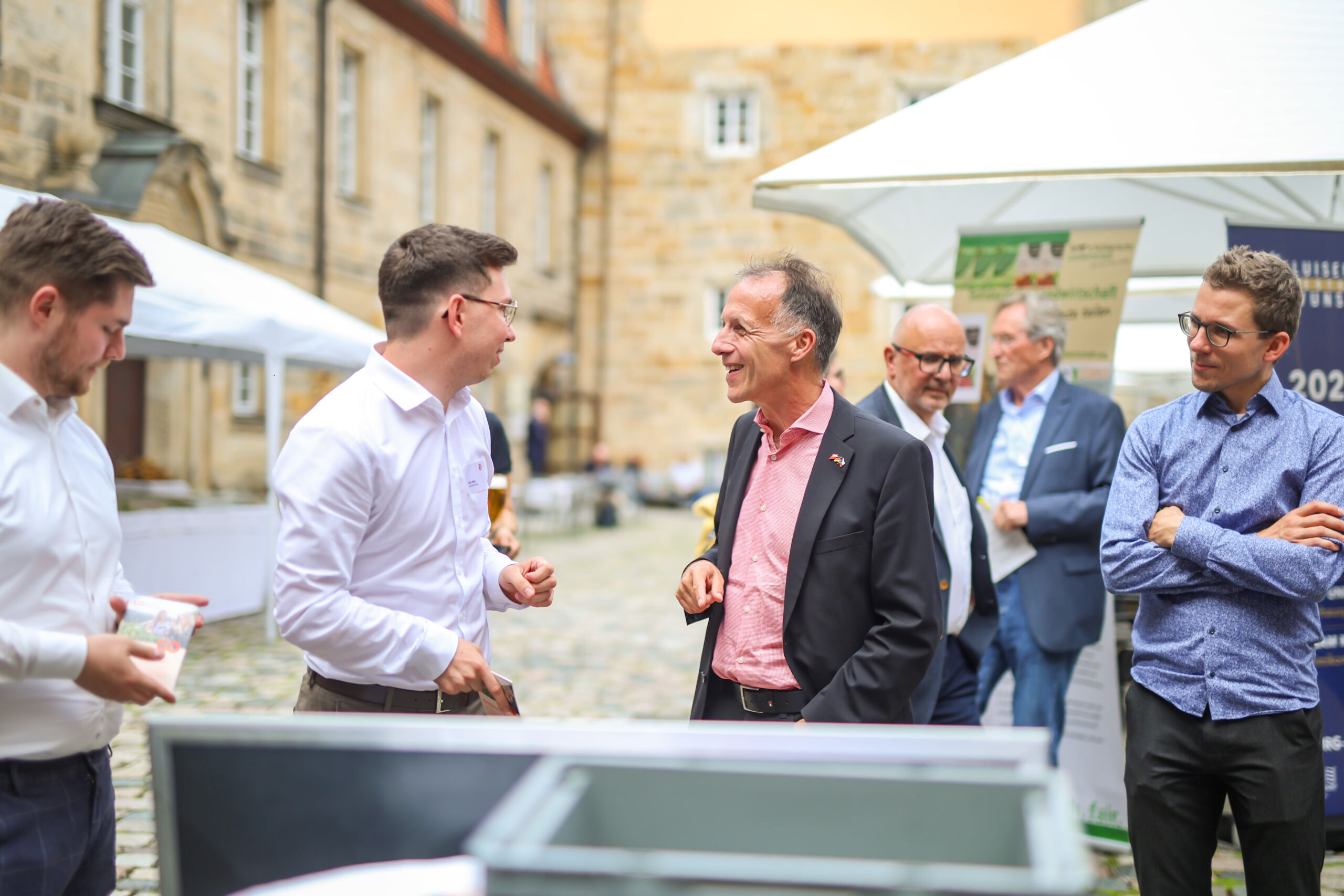 Eine Gruppe von Männern in Geschäftskleidung steht im Freien auf einem gepflasterten Innenhof des Innovationskongresses Oberfranken und unterhält sich, umgeben von alten Gebäuden und weißen Zelten; sie wirken engagiert und gut gelaunt. at Hochschule Coburg