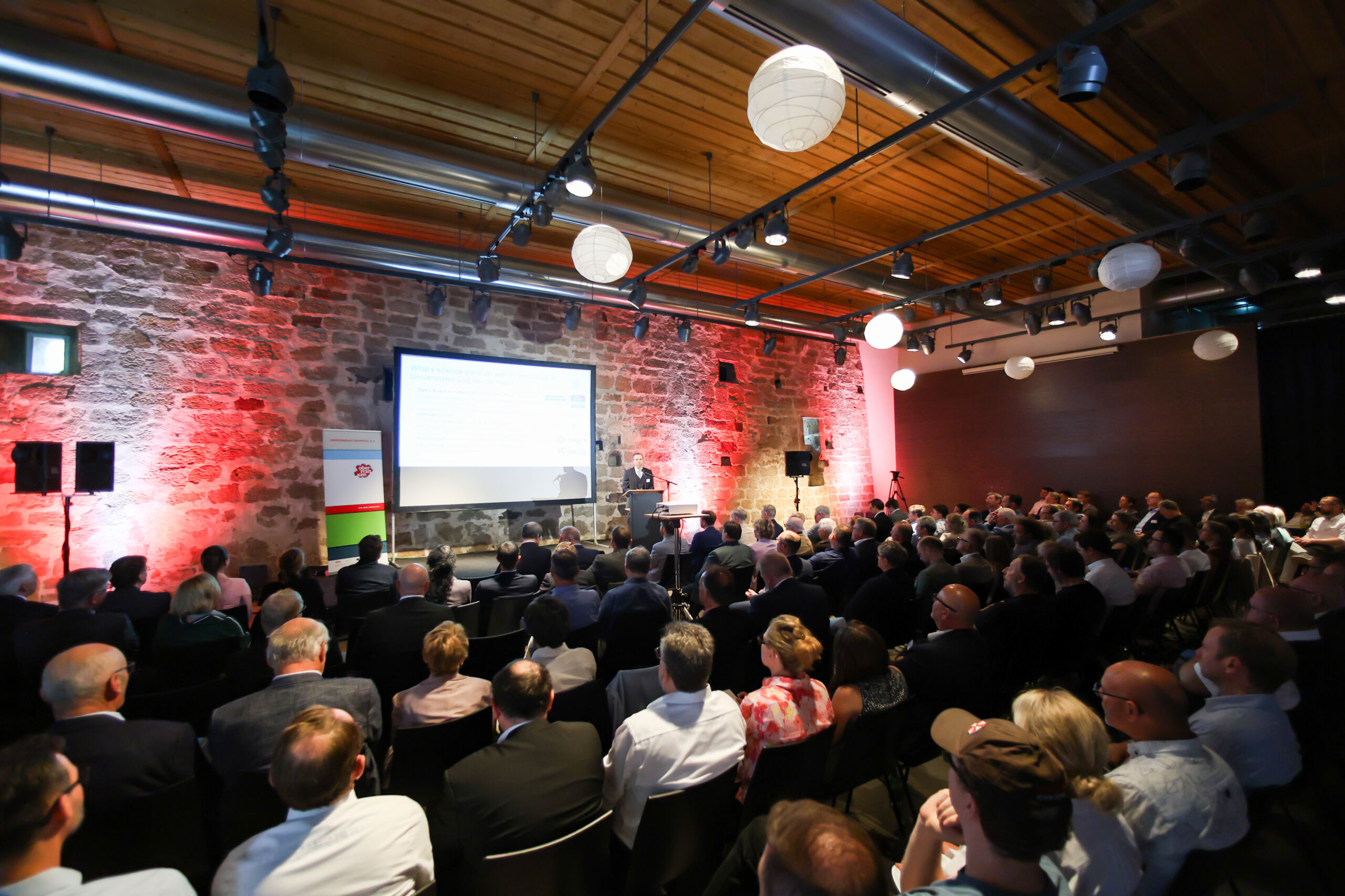 Auf dem Innovationskongress in Oberfranken hält ein Referent vor einem großen Publikum, das in einem modernen Raum mit unverputzten Ziegelwänden, Holzdecke und hängenden Leuchten Platz genommen hat, einen Vortrag über Wirtschaftsinformatik mit einer Präsentation. Das Publikum blickt aufmerksam zur Bühne. at Hochschule Coburg