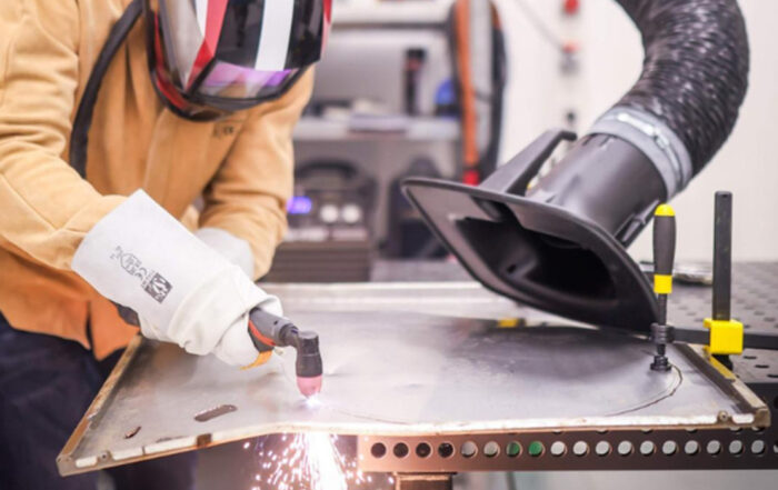 Eine Person in Schutzkleidung bearbeitet mit einem Schweißbrenner ein Blech, wobei Funken entstehen. In der Nähe ist ein Absaugschlauch angebracht, um die Dämpfe aufzufangen – ein industrieller Arbeitsbereich, in dem ZukunftsDesign in Kronach jedes Experiment inspiriert. at Hochschule Coburg