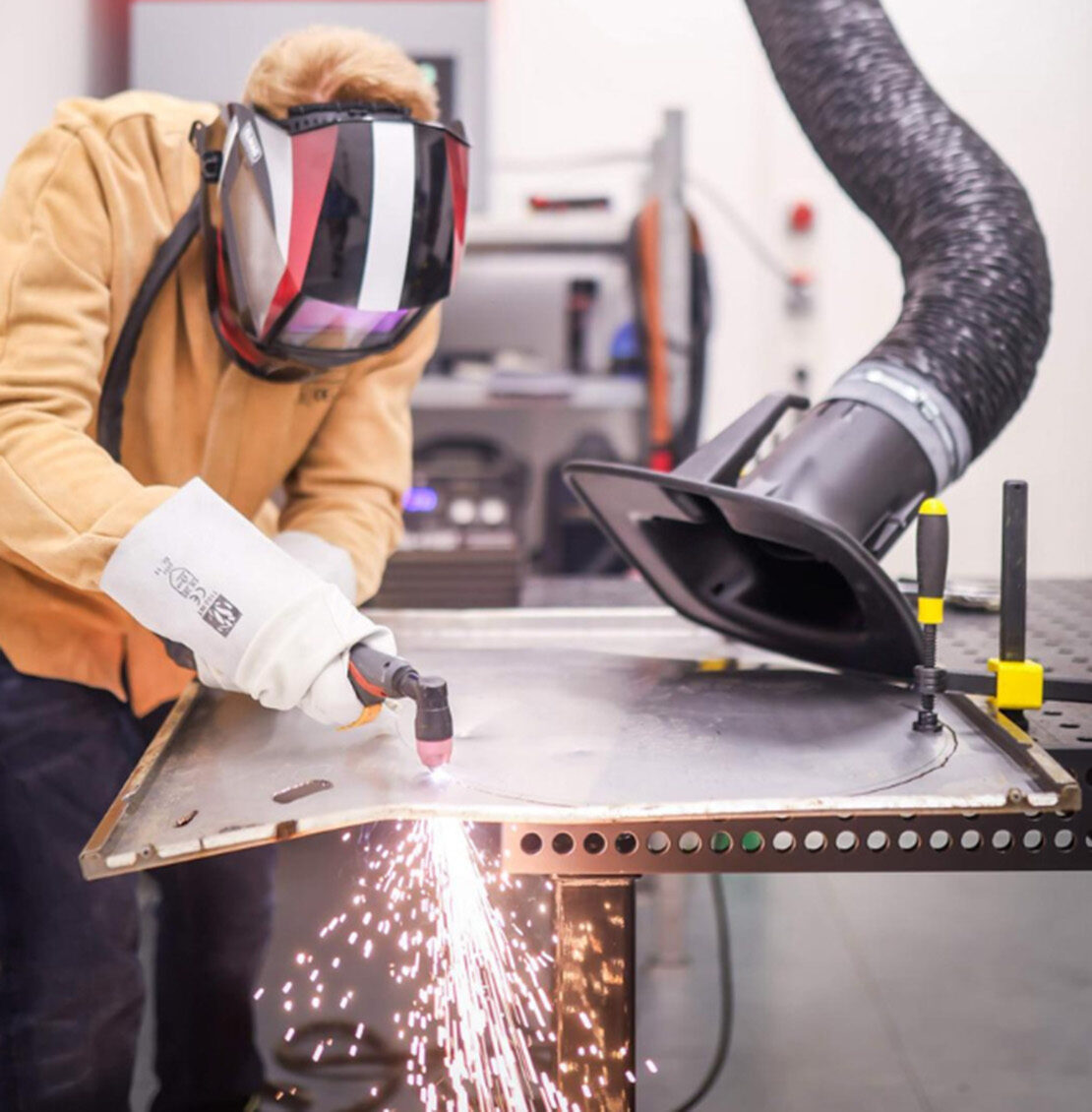 Eine Person in Schutzkleidung bearbeitet mit einem Schweißbrenner ein Blech, wobei Funken entstehen. In der Nähe ist ein Absaugschlauch angebracht, um die Dämpfe aufzufangen – ein industrieller Arbeitsbereich, in dem ZukunftsDesign in Kronach jedes Experiment inspiriert. at Hochschule Coburg