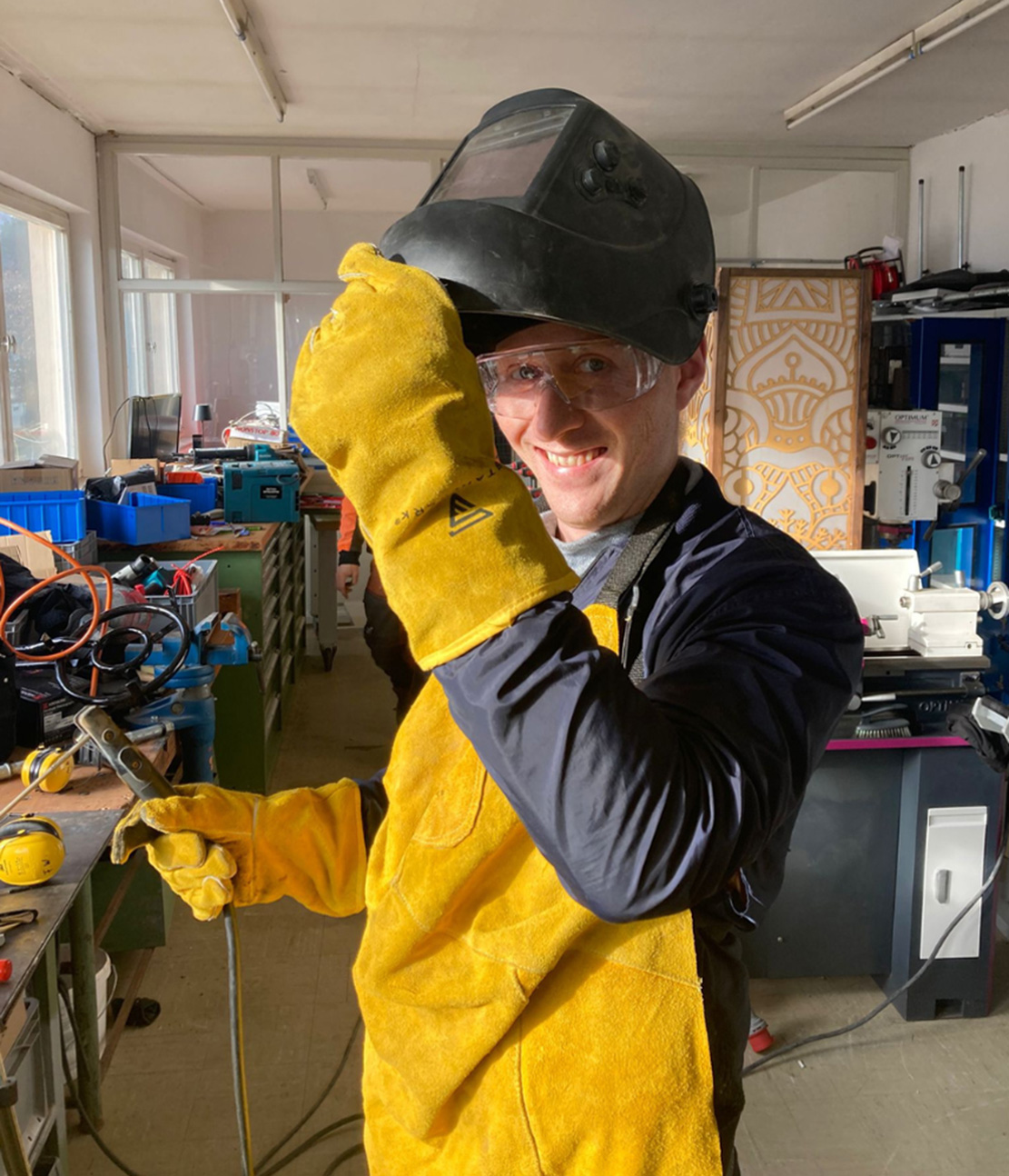 Eine Person in einer Werkstatt in Kronach trägt einen Schweißhelm, gelbe Handschuhe und eine Schutzschürze und lächelt, während sie den Helm hochhebt. Umgeben von Werkzeugen und Maschinen verkörpert sie den Geist von Experiment und ZukunftsDesign. at Hochschule Coburg