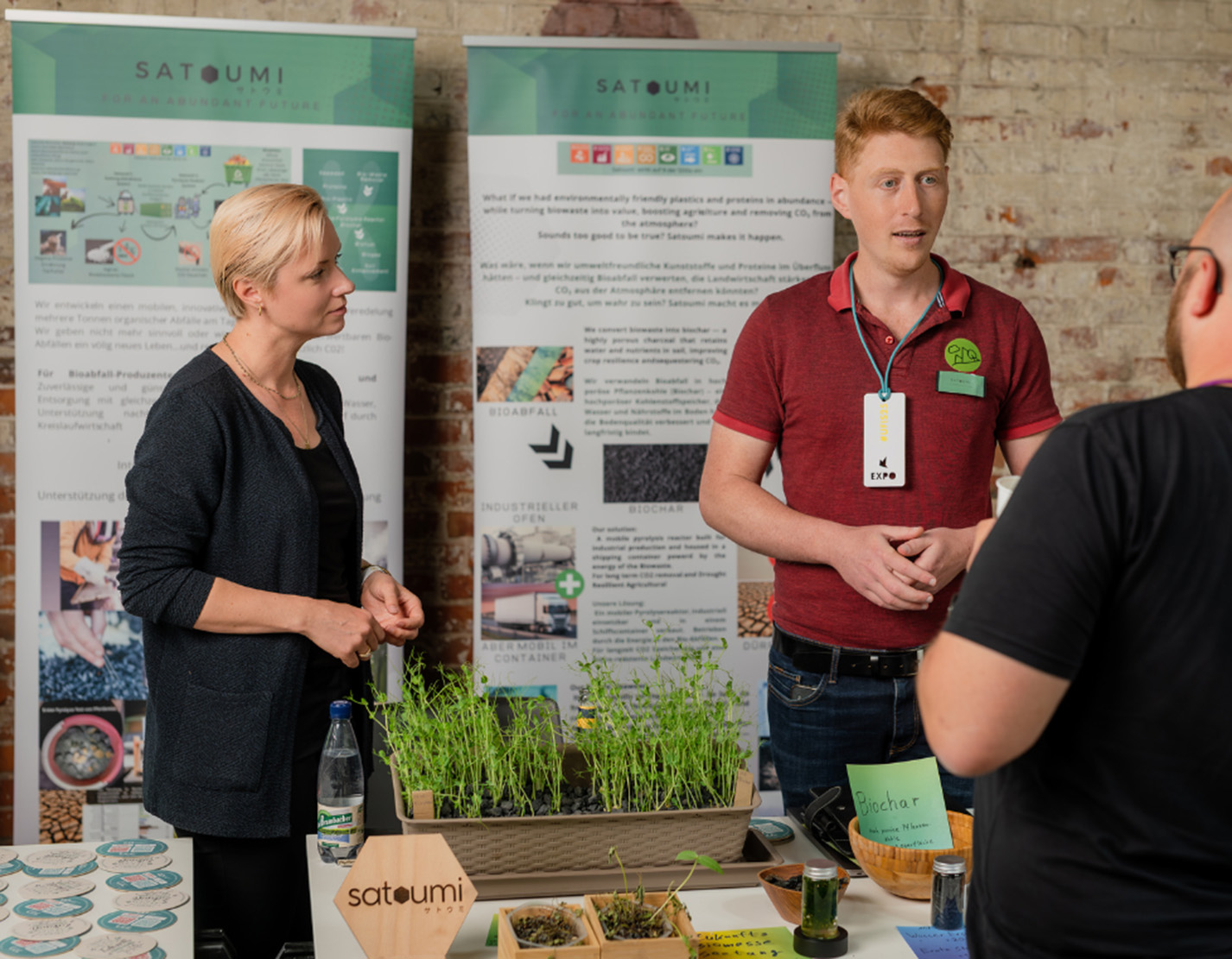 Zwei Personen an einem Stand mit Informationsplakaten, Pflanzen und Mustern sprechen bei einer Indoor-Veranstaltung zum Thema Nachhaltigkeit in Kronach mit einem Besucher und stellen dabei innovative Ideen im Bereich ZukunftsDesign sowie experimentelle Ansätze vor. at Hochschule Coburg