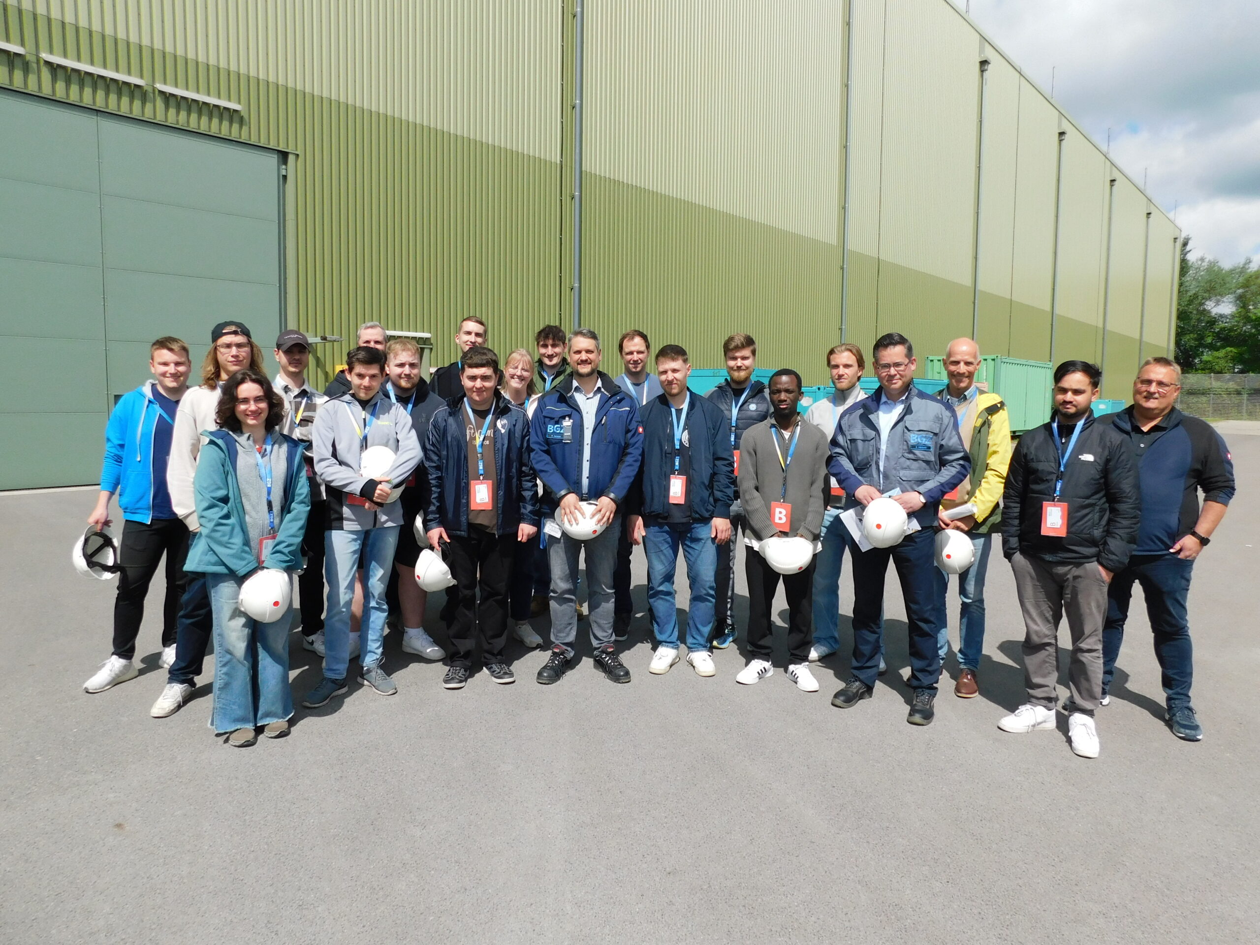 Eine Gruppe von Menschen, darunter auch Coburger Studierende, einige mit weißen Schutzhelmen, steht an einem sonnigen Tag vor einem großen grünen Industriegebäude in Grafenrheinfeld zusammen. Die meisten tragen Jacken und haben Namensschilder oder Schlüsselbänder - möglicherweise diskutieren sie über Atommüll. at Hochschule Coburg