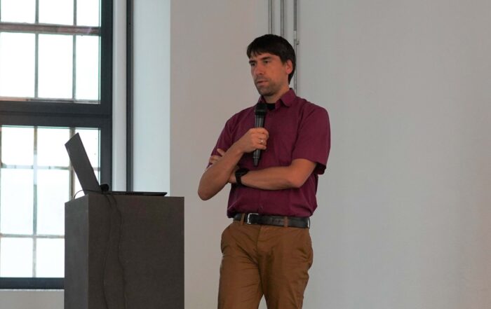 Ein Mann in einem kastanienbraunen Hemd und einer braunen Hose hält ein Mikrofon in der Nähe eines Podiums mit einem Laptop. Er spricht bei einem Themenabend über moderne Krebstherapien in einem hellen Raum mit großen Fenstern. at Hochschule Coburg