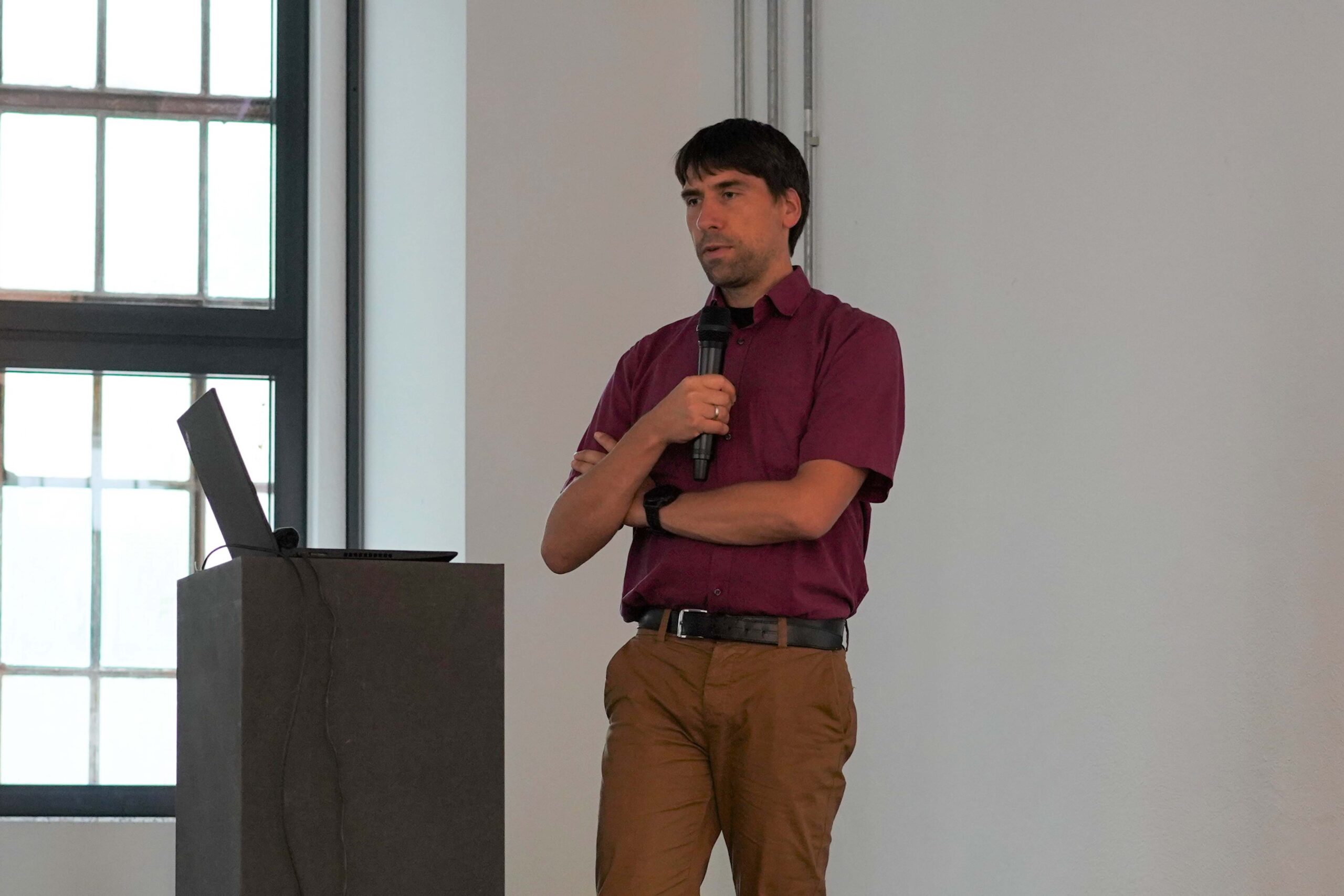 Ein Mann in einem kastanienbraunen Hemd und einer braunen Hose hält ein Mikrofon in der Nähe eines Podiums mit einem Laptop. Er spricht bei einem Themenabend über moderne Krebstherapien in einem hellen Raum mit großen Fenstern. at Hochschule Coburg