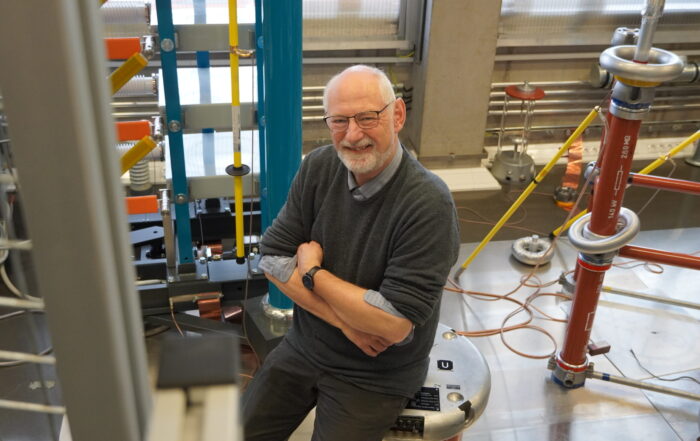 Ein älterer Mann mit Brille und Bart steht lächelnd in einem Labor, das mit hohen elektrotechnischen Geräten, Kabeln und Metalloberflächen gefüllt ist; er trägt einen grauen Pullover und ein blaues Hemd – und verkörpert Wandel in seiner Rolle als Professor. at Hochschule Coburg