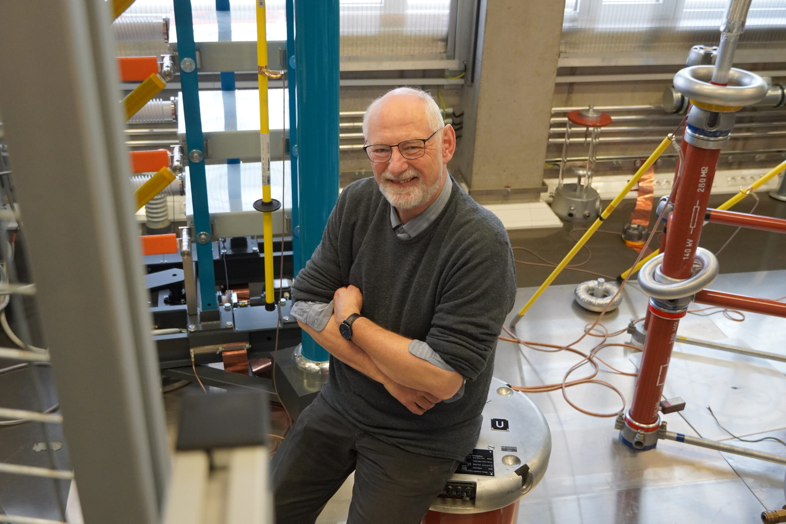 Ein älterer Mann mit Brille und Bart steht lächelnd in einem Labor, das mit hohen elektrotechnischen Geräten, Kabeln und Metalloberflächen gefüllt ist; er trägt einen grauen Pullover und ein blaues Hemd – und verkörpert Wandel in seiner Rolle als Professor. at Hochschule Coburg