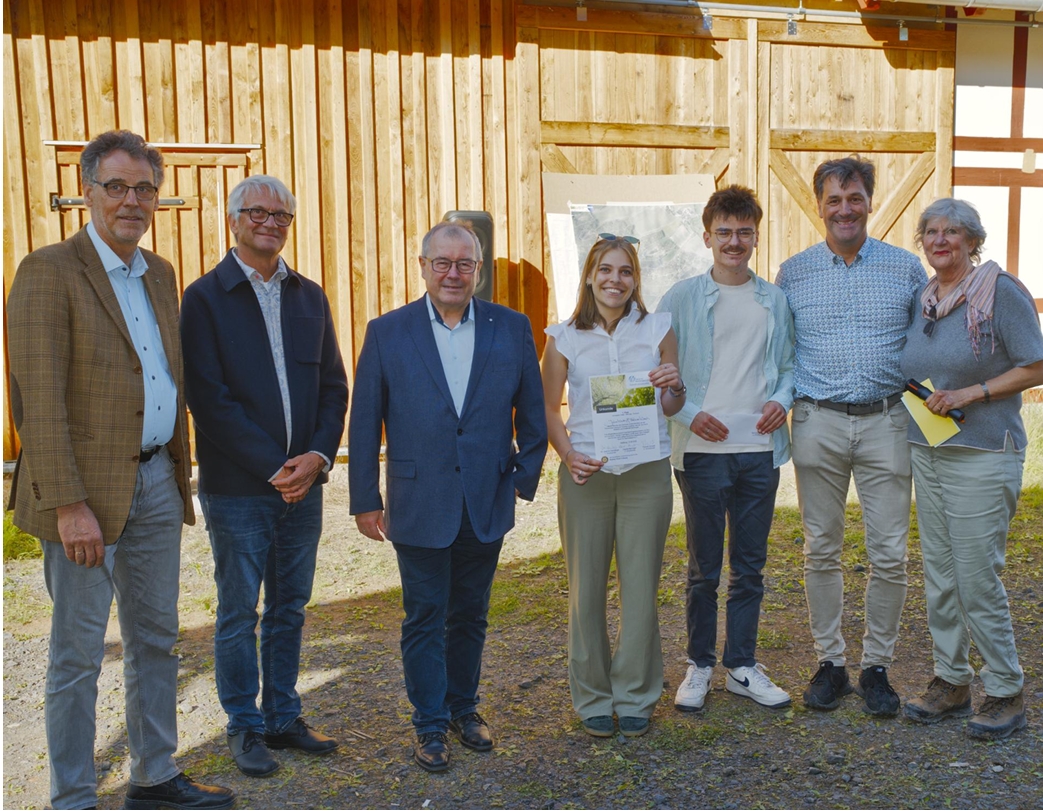 Sieben Menschen stehen lächelnd im Freien vor einem Holzgebäude am Bildungszentrum Einöd; zwei junge Leute in der Mitte halten ein Zertifikat in den Händen, umgeben von anderen in legerer Kleidung, im Hintergrund ist der grüne Wald zu sehen. at Hochschule Coburg