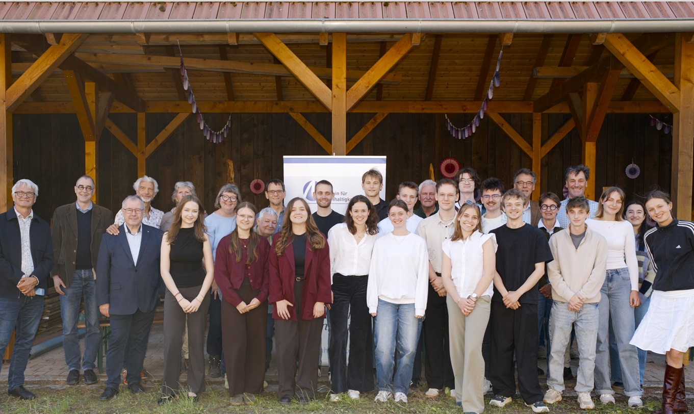Eine große Gruppe von Menschen, darunter Erwachsene und junge Erwachsene, steht lächelnd vor einem Holzpavillon im Bildungszentrum Einöd, hinter ihnen ein Transparent, und posiert an einem sonnigen Tag im Freien am Waldrand für ein Gruppenfoto. at Hochschule Coburg