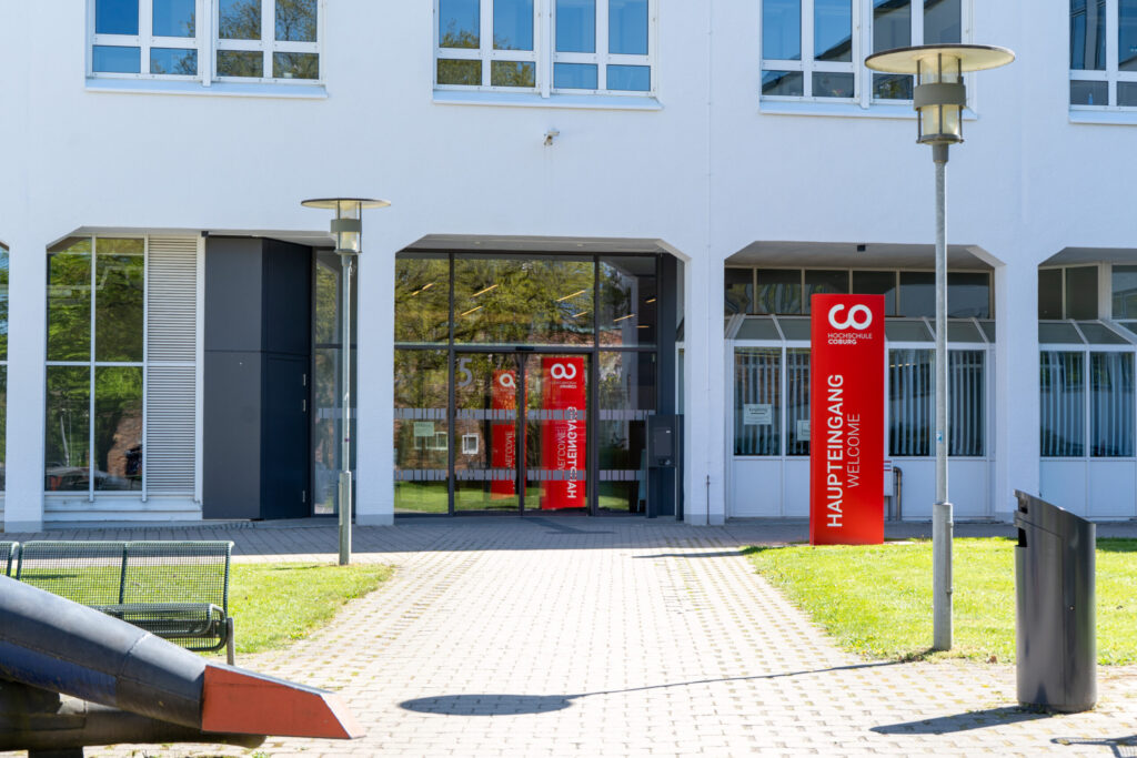 Das Bild zeigt den Haupteingang eines modernen weißen Gebäudes mit großen Fenstern. Auf roten Schildern neben der Tür steht "HAUPTEINGANG WILLKOMMEN". Es gibt Bänke, Straßenlaternen und einen Weg, der zum Eingang führt. at Hochschule Coburg