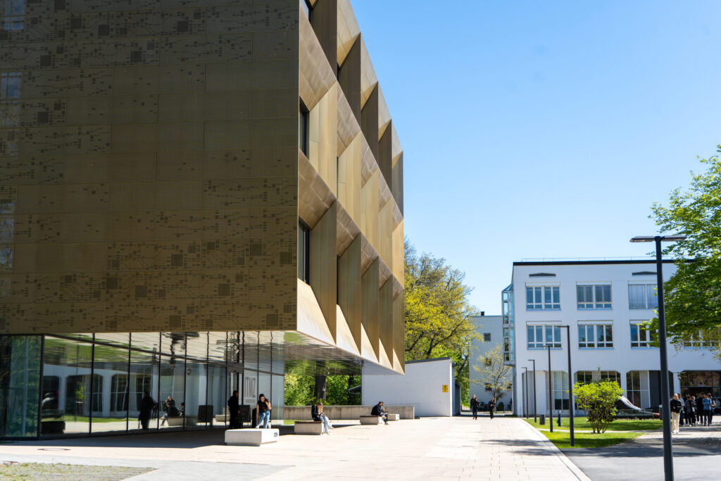 Ein modernes, goldfarbenes Gebäude mit eckigen Fenstern und einem Glassockel steht an einem sonnigen Tag neben einem weißen, rechteckigen Gebäude; draußen sitzen und gehen Menschen auf einem gepflasterten Gehweg, der von grünen Rasenflächen und Bäumen umgeben ist. at Hochschule Coburg