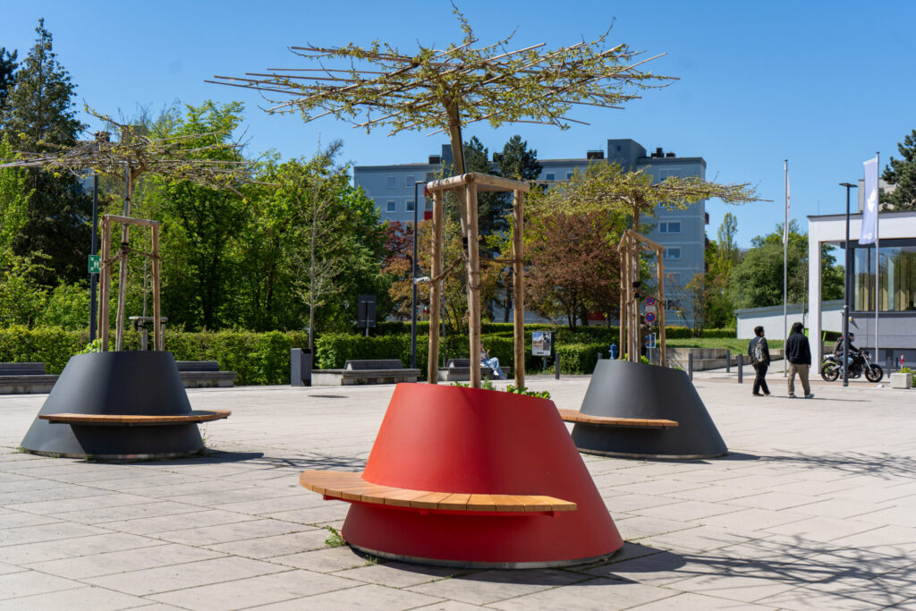Moderne öffentliche Bänke mit eingebauten Pflanzgefäßen und jungen Bäumen auf einem sonnenbeschienenen städtischen Platz, umgeben von Grünflächen, Gebäuden und einigen Spaziergängern im Hintergrund. at Hochschule Coburg
