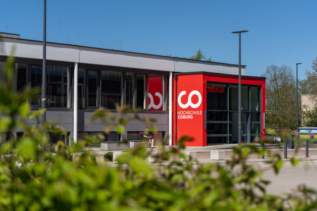 Ein modernes Gebäude mit großen Fenstern und einem auffälligen roten Schild mit der Aufschrift "Hochschule Coburg" in der Nähe des Eingangs, das teilweise durch grünes Laub im Vordergrund verdeckt wird. at Hochschule Coburg